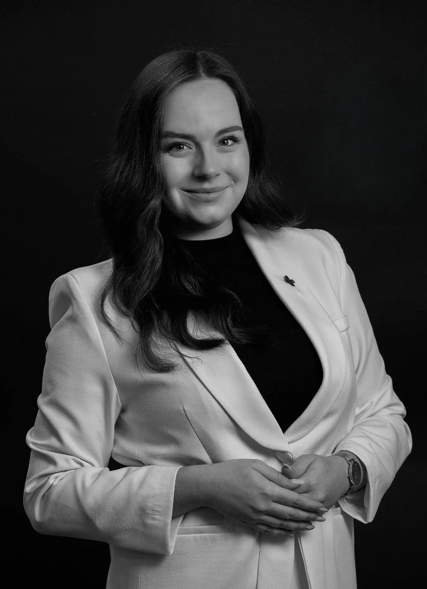 Black and white portrait of a young woman with dark, wavy hair, wearing a light-colored blazer over a dark top, smiling softly at the camera