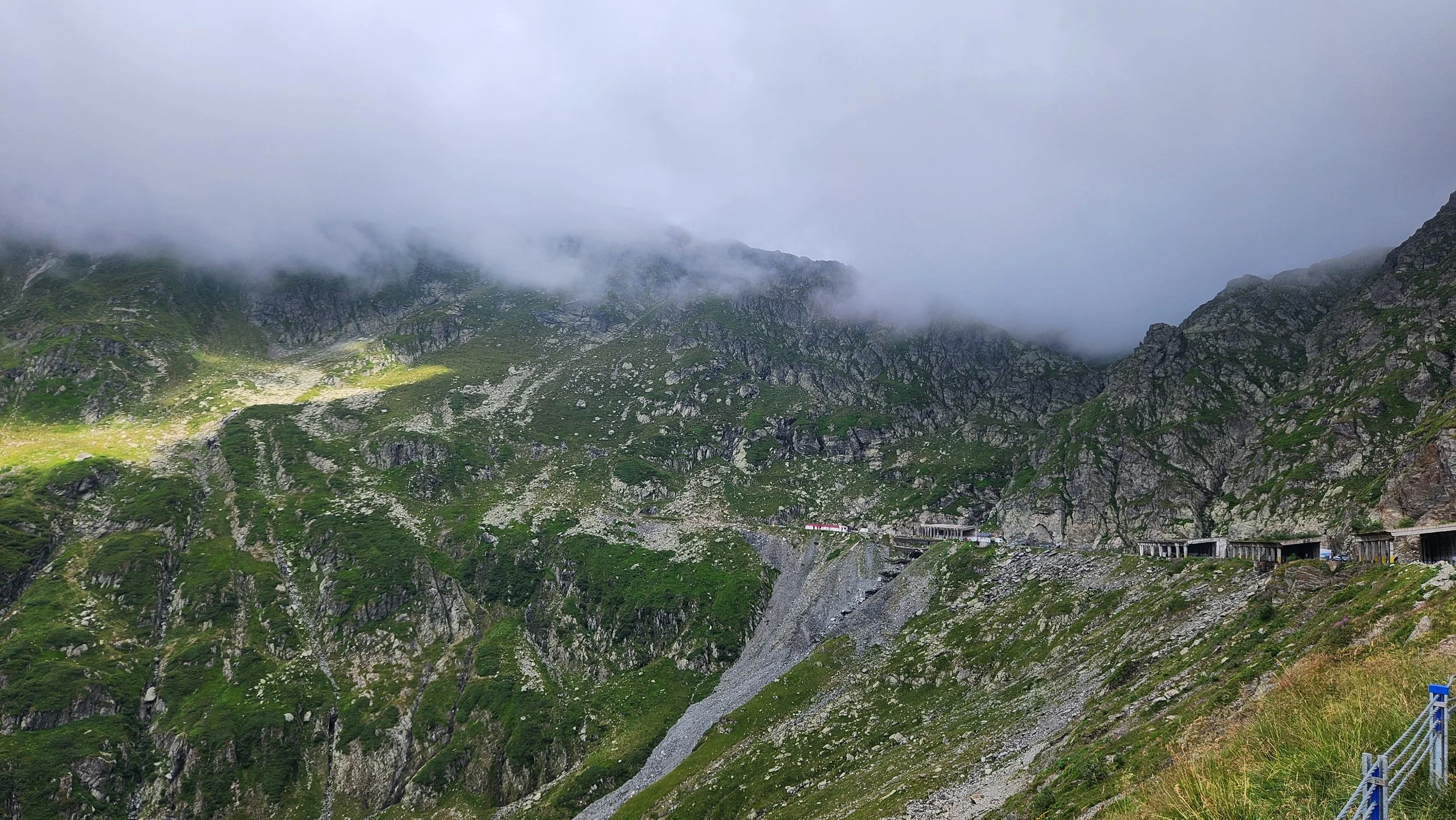 Berglandschaft mit grünen Hängen, Felsen und Nebel, darunter eine Bahnstrecke mit Tunneln