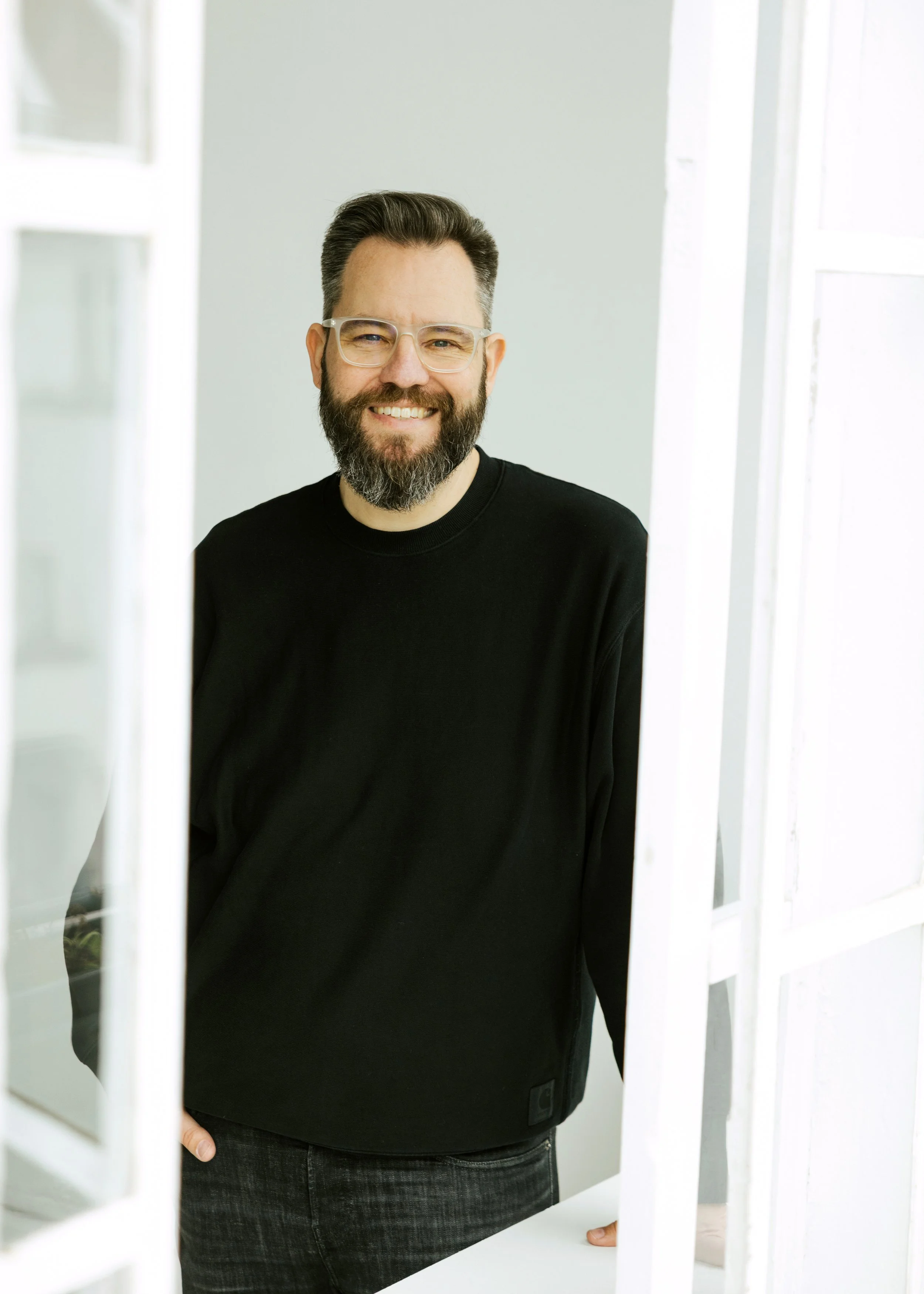 Ein lachender Mann mit Bart und Brille, trägt ein schwarzes Sweatshirt, steht in einem hellen Raum, teilweise sichtbar durch eine weiße Fensterrahmen