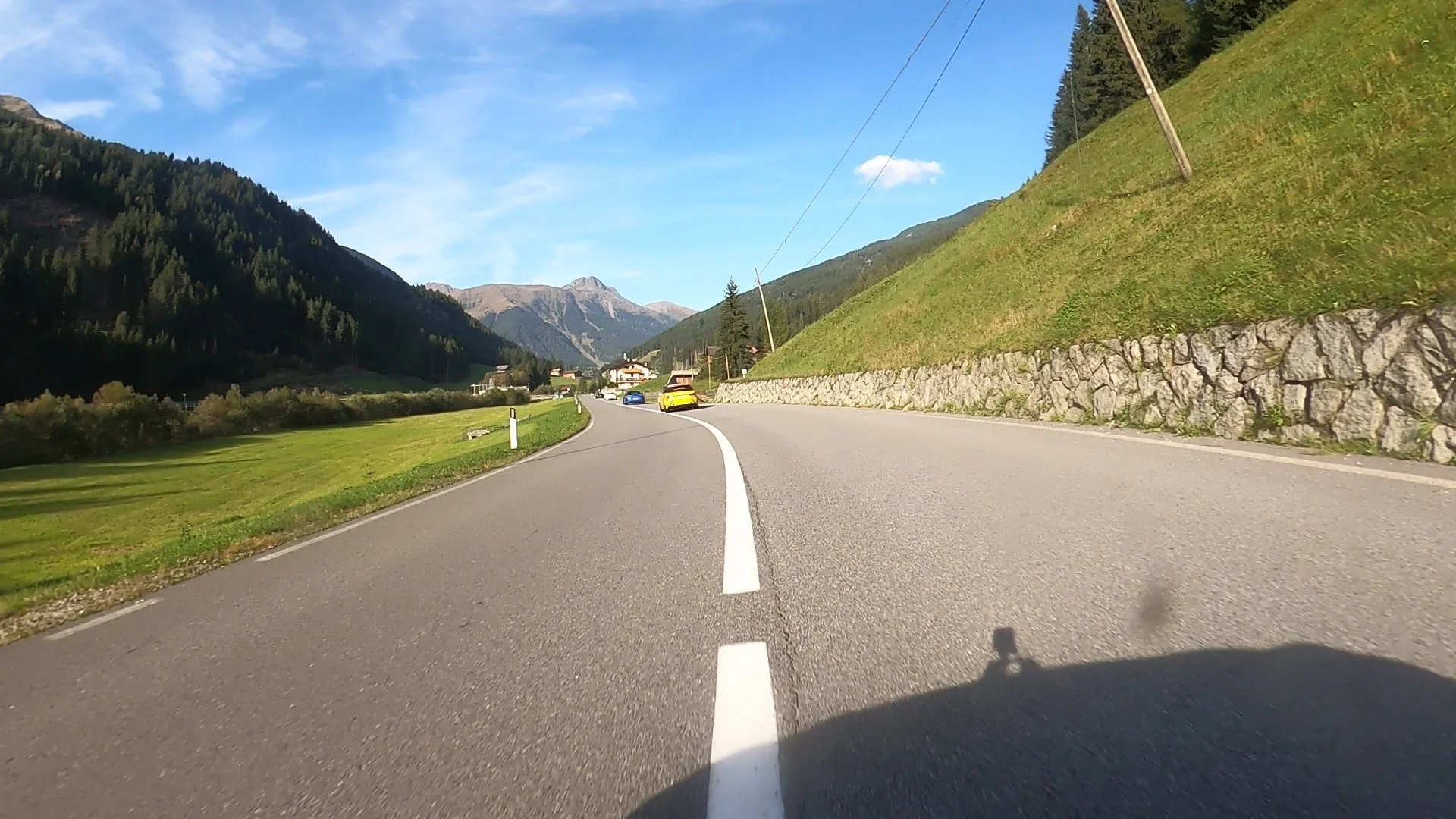 Blick auf eine kurvige Landstraße im Gebirge mit mehreren Autos, grünen Hügeln, Bergen im Hintergrund und blauer Himmel