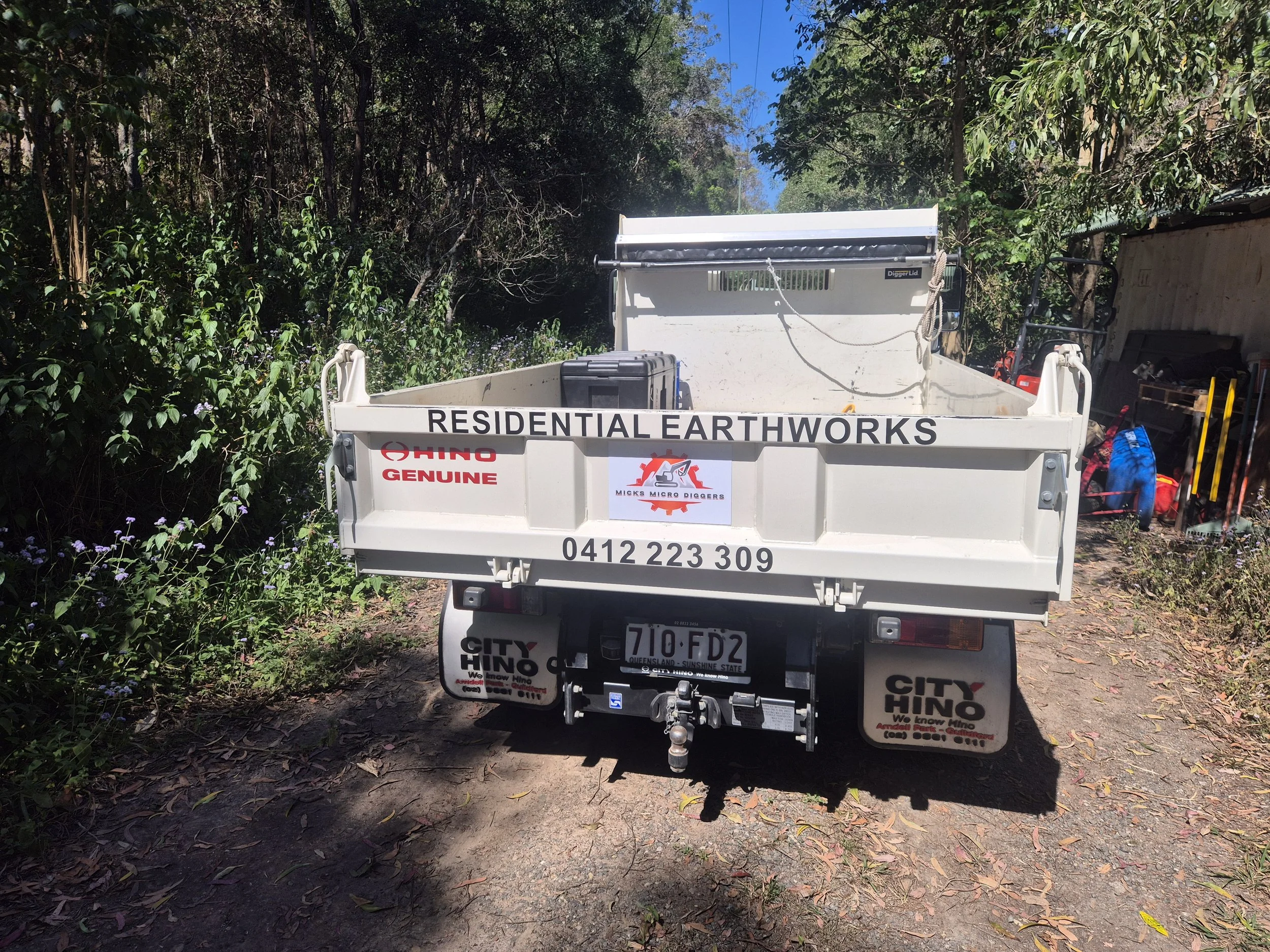 Pickup truck with a sign that reads 'RESIDENTIAL EARTHWORKS' on the back, parked on a dirt path surrounded by greenery and trees.