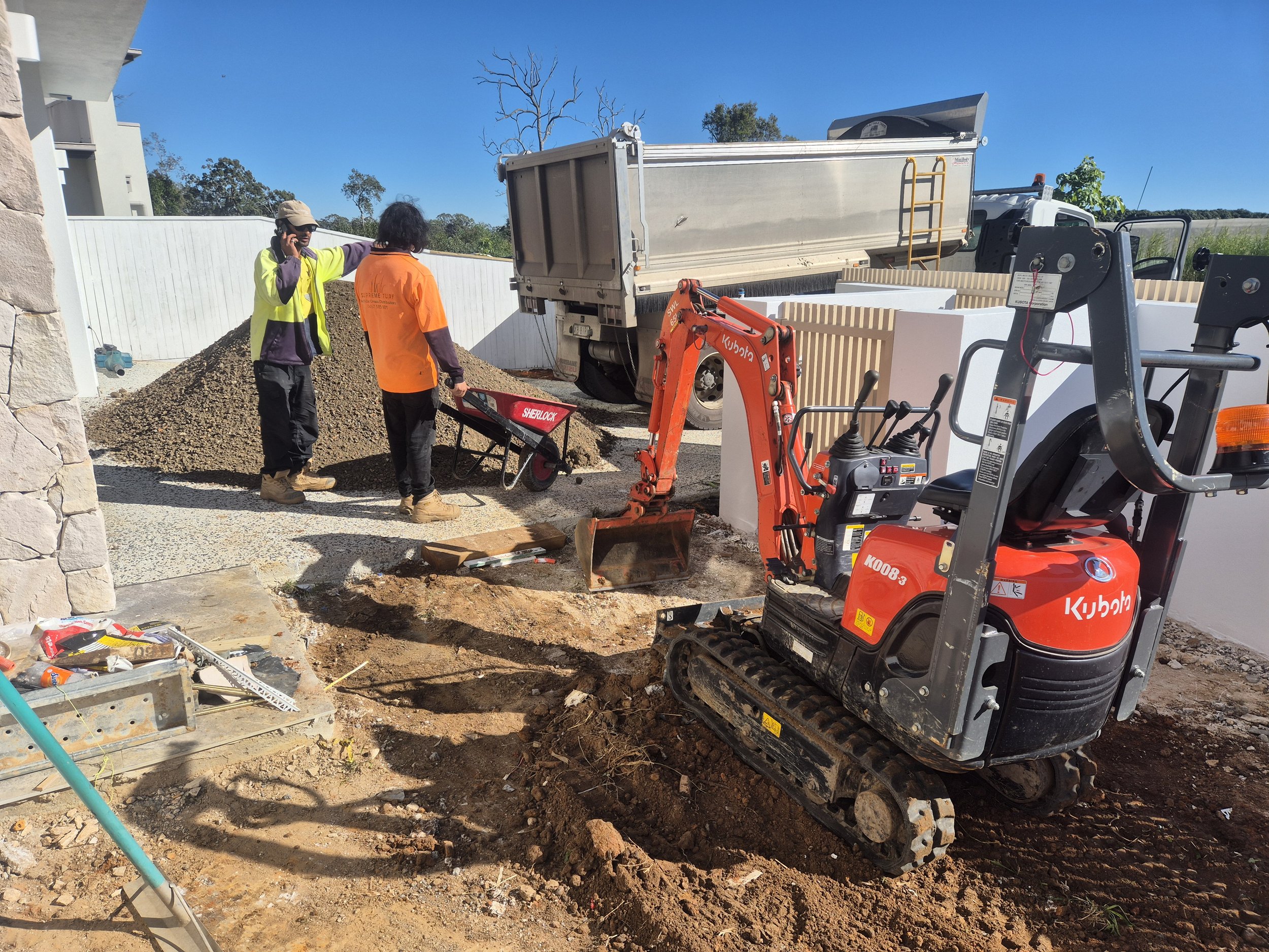 Construction workers and equipment working on a construction site near a house, with a mini excavator, a wheelbarrow, and a dump truck under a clear blue sky.