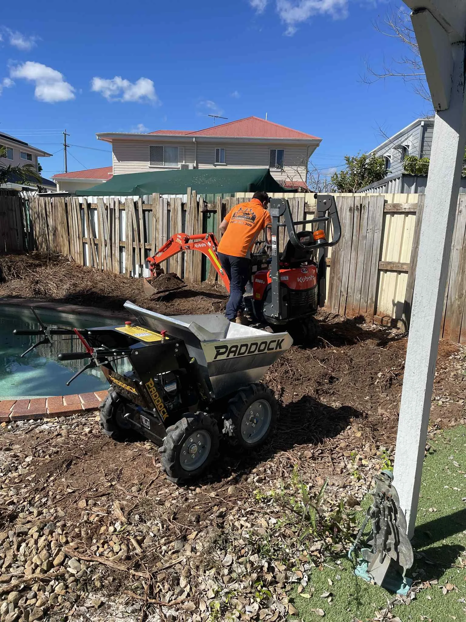 A person operating a small Kubota excavator digging near a wooden fence in a backyard with a pond. There is a small Paddock landscape rake on the ground. The sky is blue with some clouds, and surrounding houses are visible.