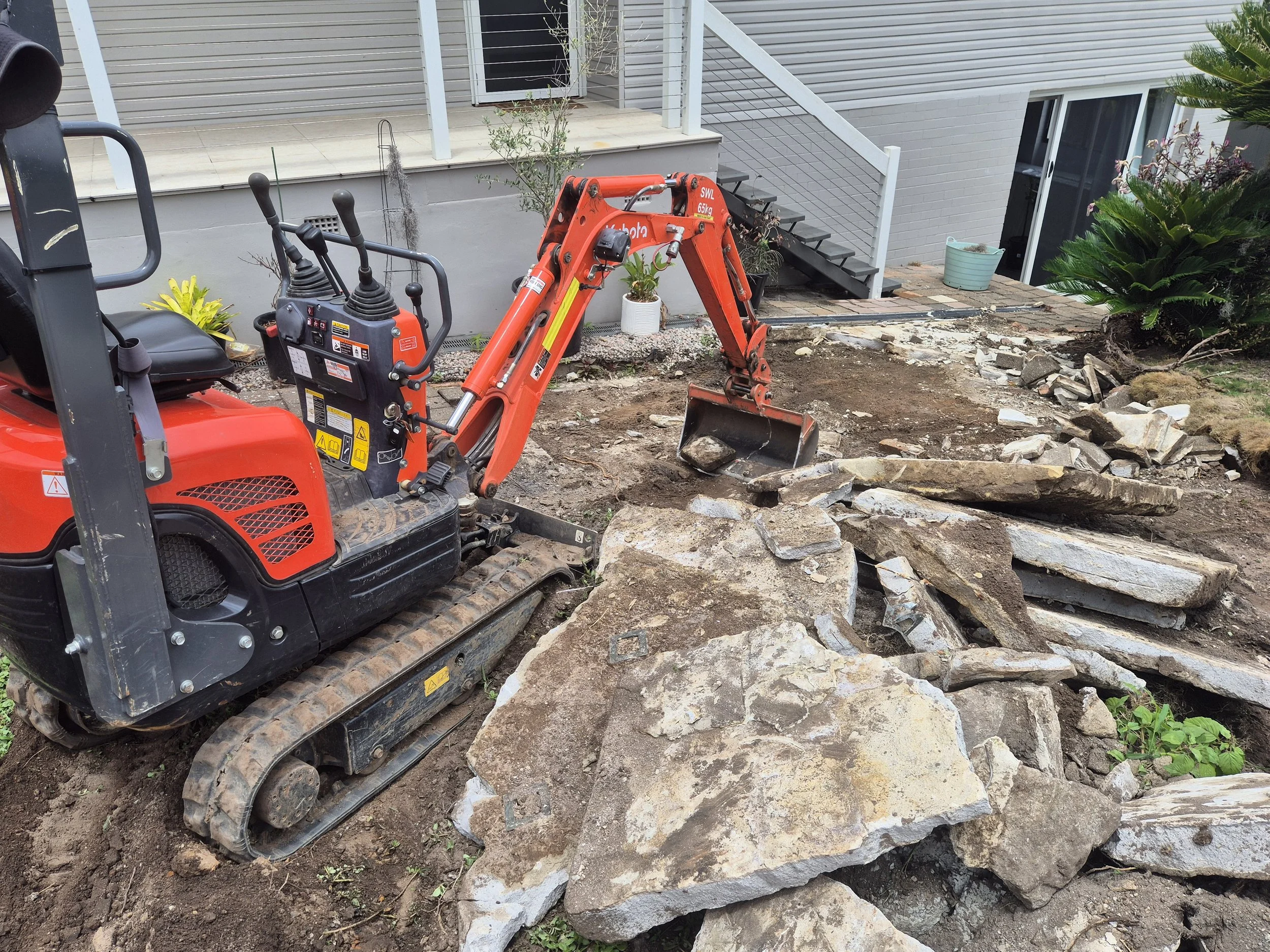 A mini excavator working on outdoor stone landscaping near a house with stairs and plants.