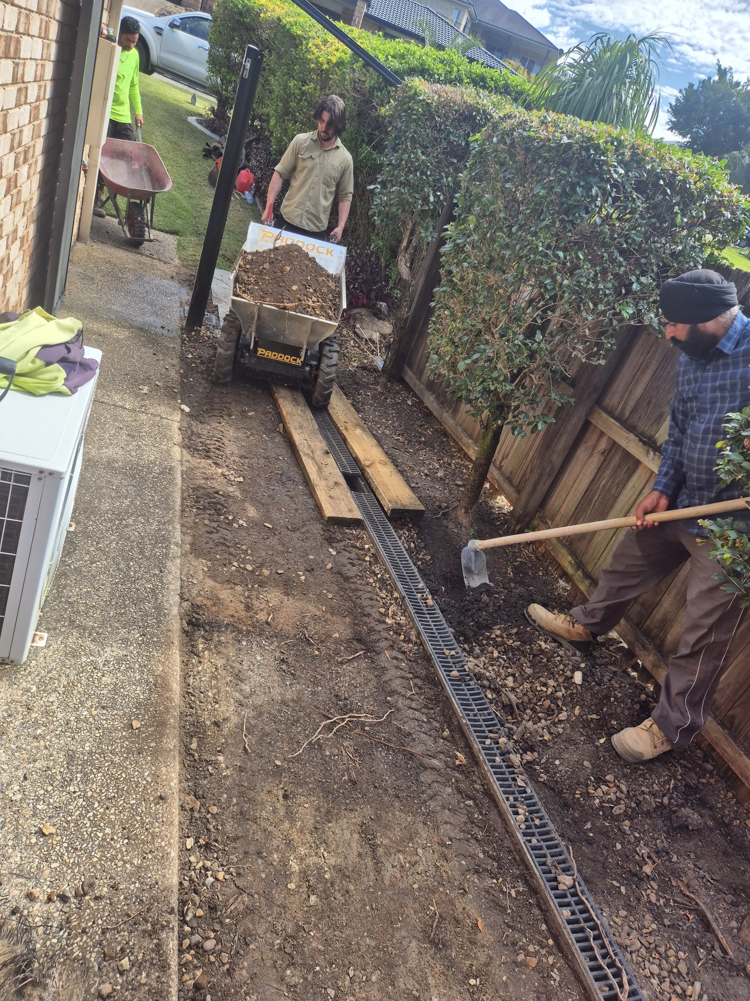 People working in a backyard installing a drainage system with gravel, a wheelbarrow, and shovels. There are trees and bushes along a wooden fence.