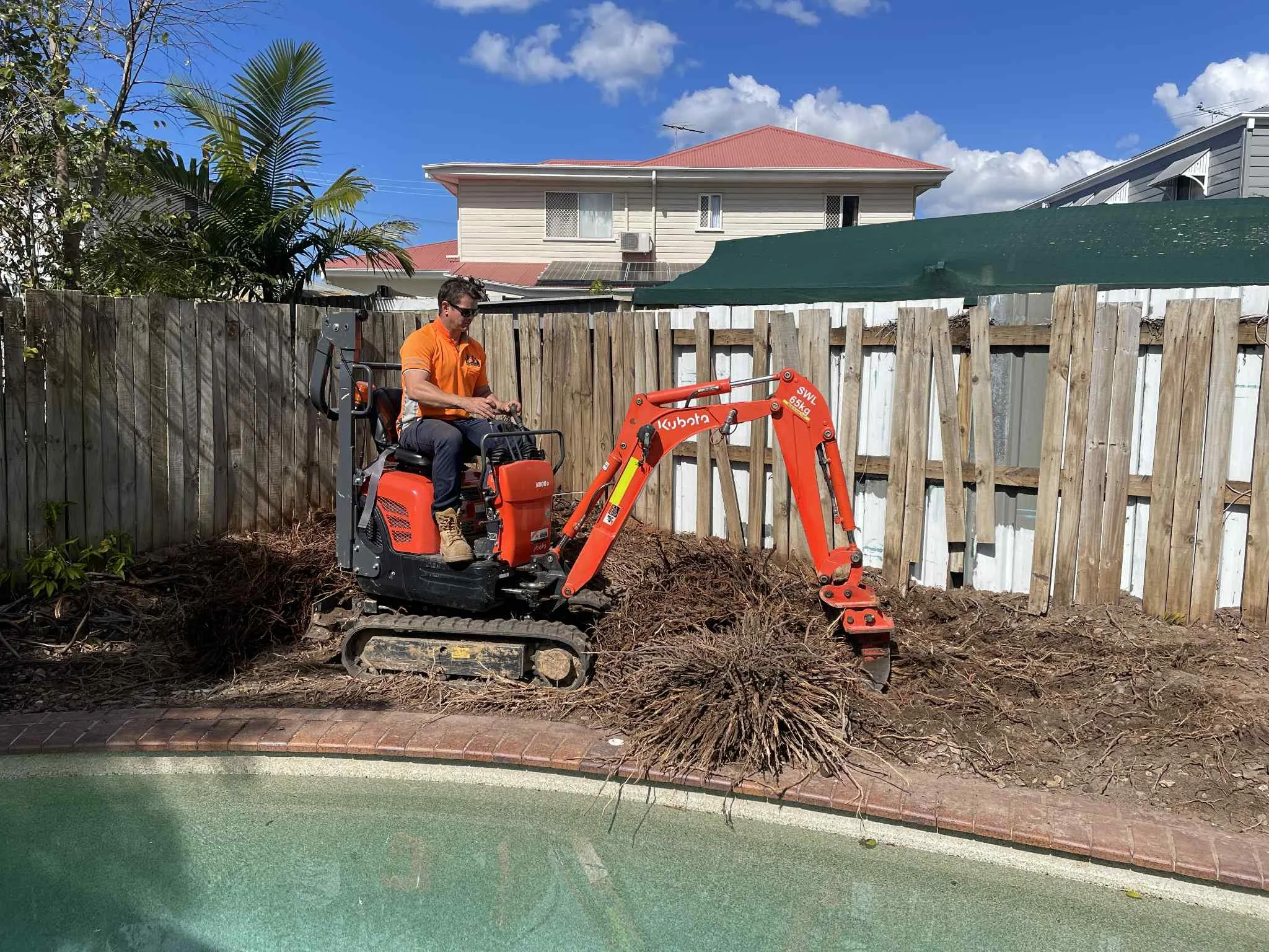 A person in an orange shirt and sunglasses operating a small orange Kubota mini excavator beside a backyard swimming pool, digging up soil near a wooden fence with two houses in the background under a blue sky.