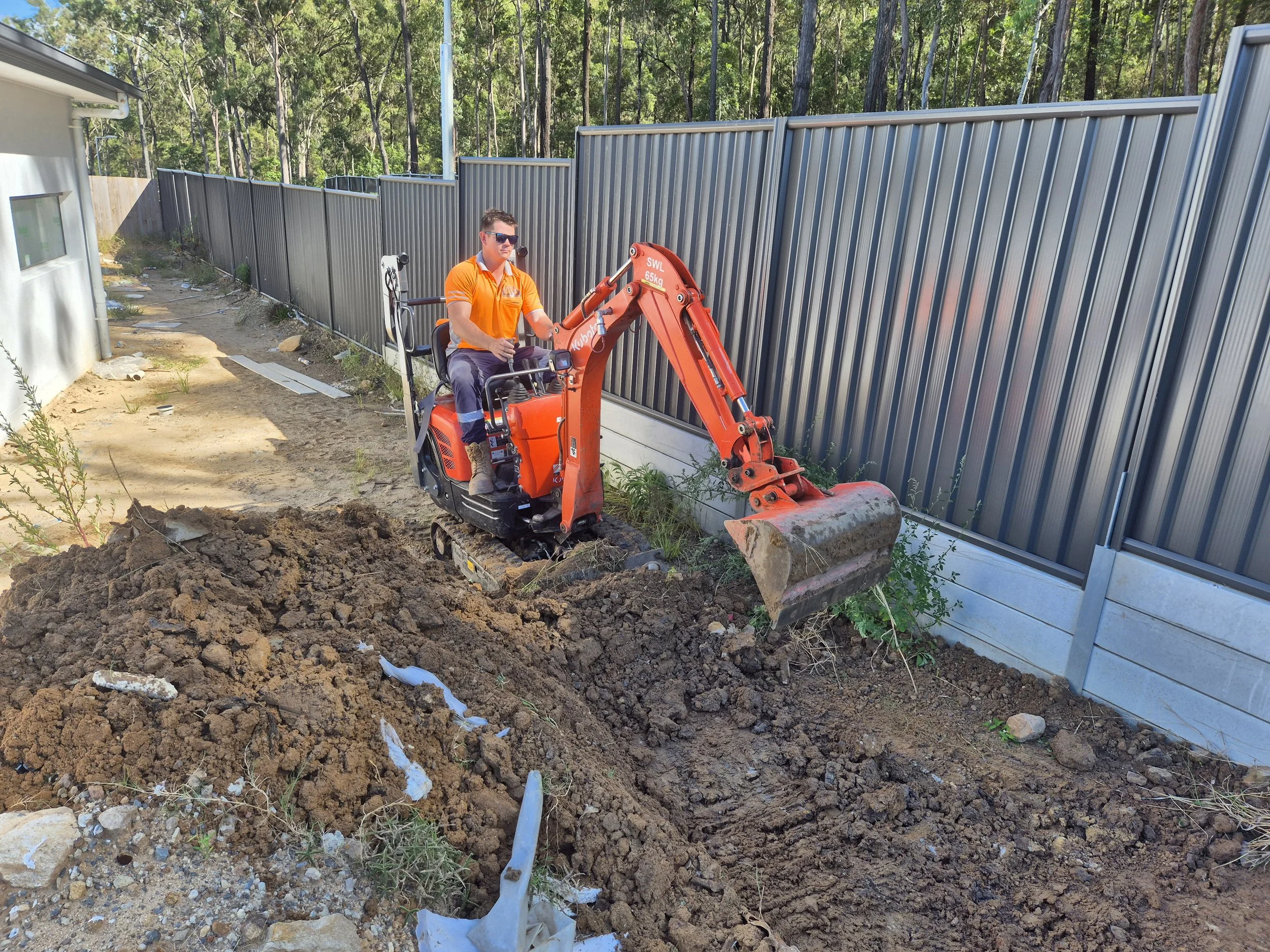 Man operating a mini excavator digging near a metal fence in a backyard.