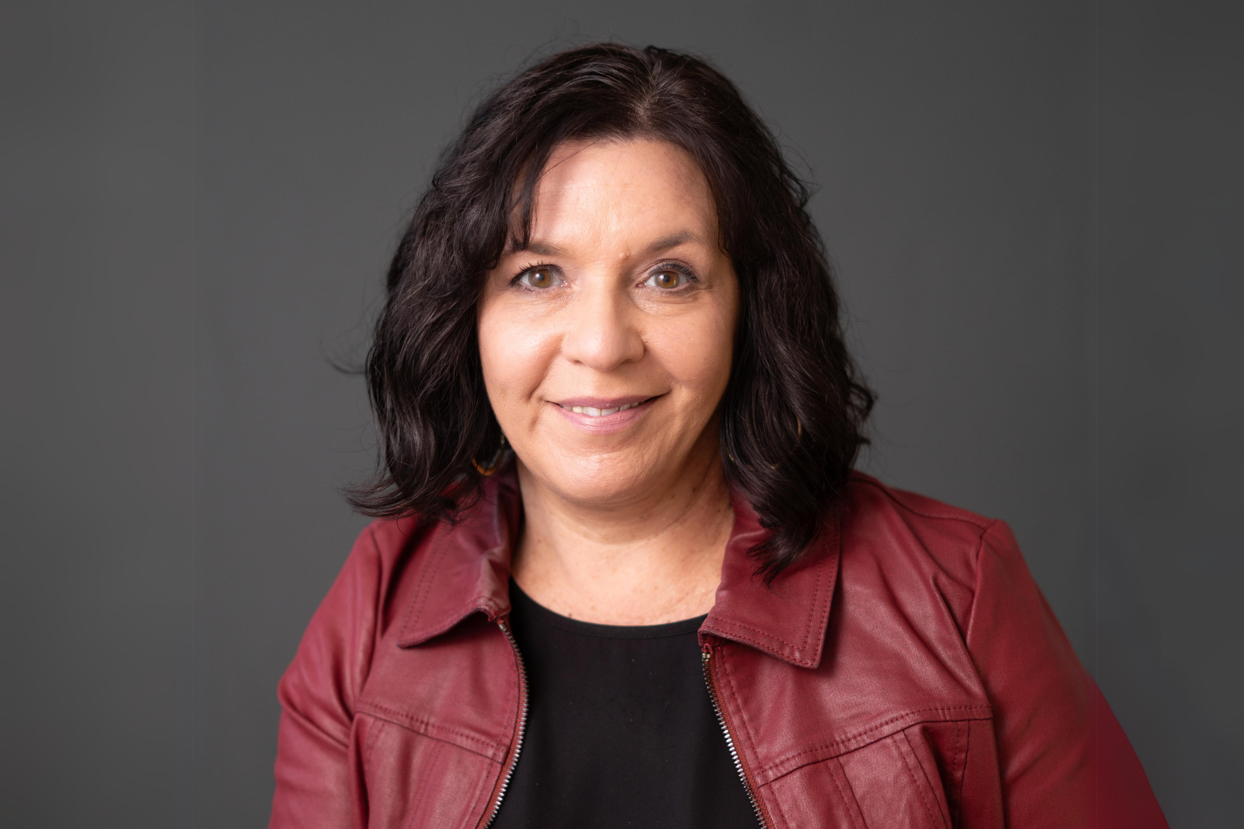 A woman with shoulder-length dark hair, wearing a red leather jacket and black top, smiling at the camera against a gray background.
