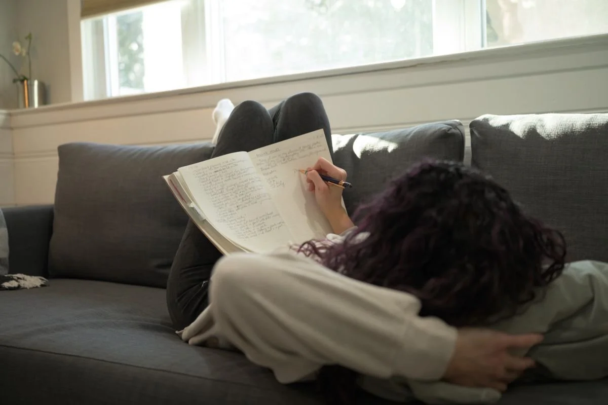 Woman journaling in a notebook on a couch in soft morning light, representing small daily steps, reflection, and building momentum in everyday life.