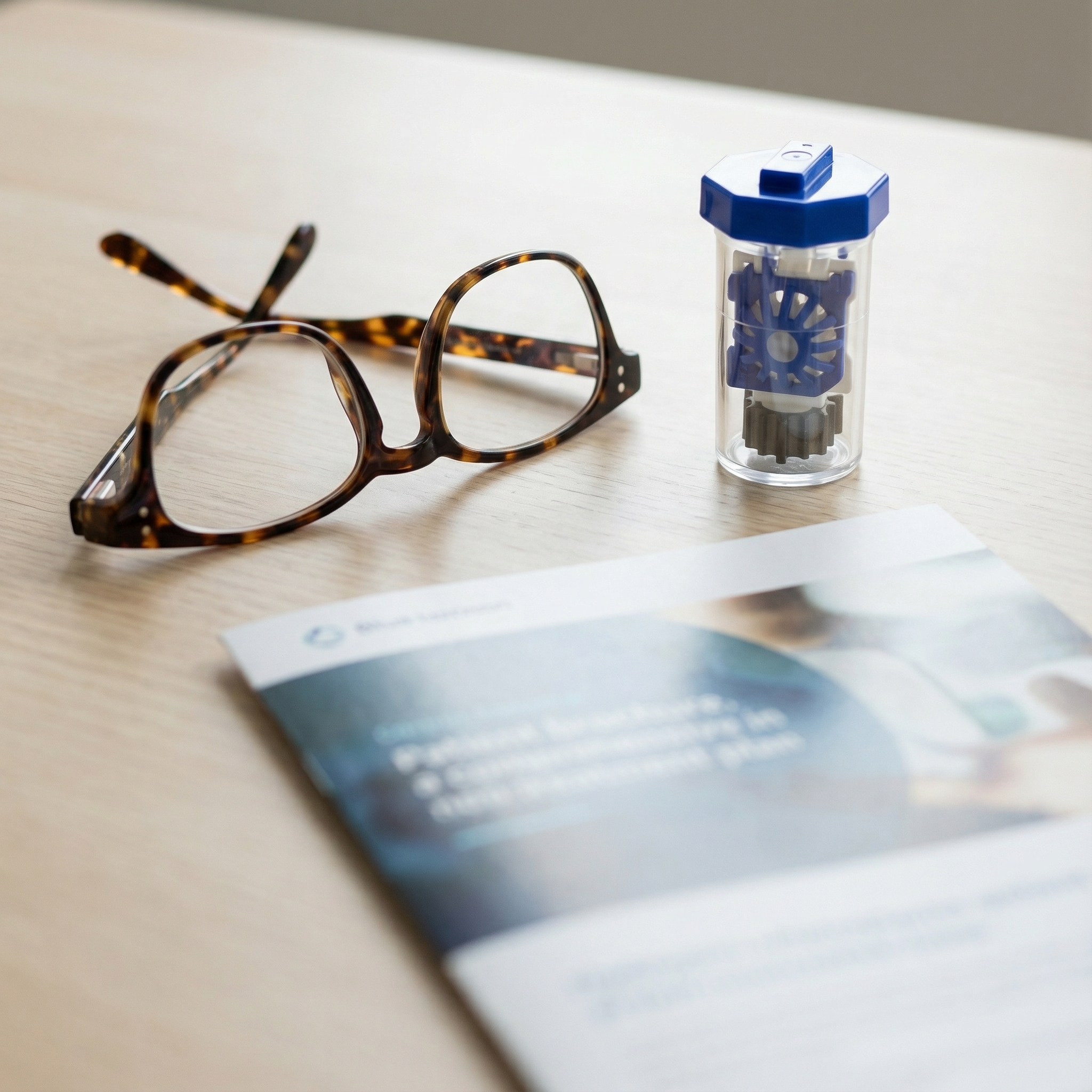 Tortoise shell eyeglasses, contact lens case with lens cleaning solution, and a blurred brochure or pamphlet on a light-colored wooden surface.