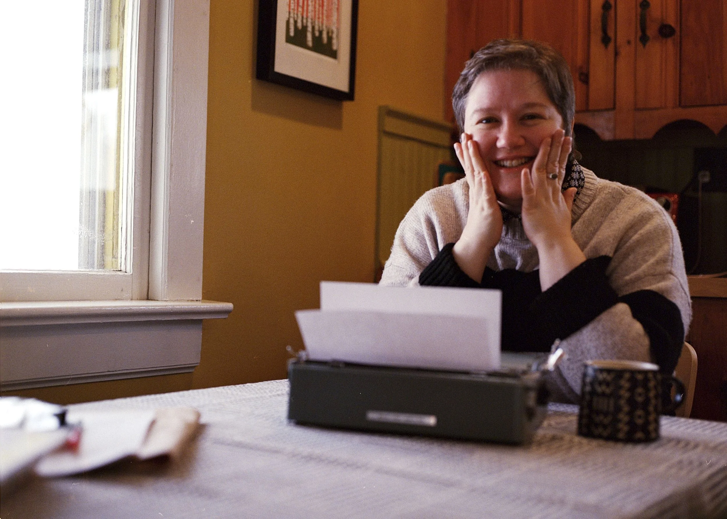 A woman with short hair, smiling and touching her face, sitting at a table with a typewriter, a coffee mug, and some papers, in a cozy room with wooden cabinets and a window.