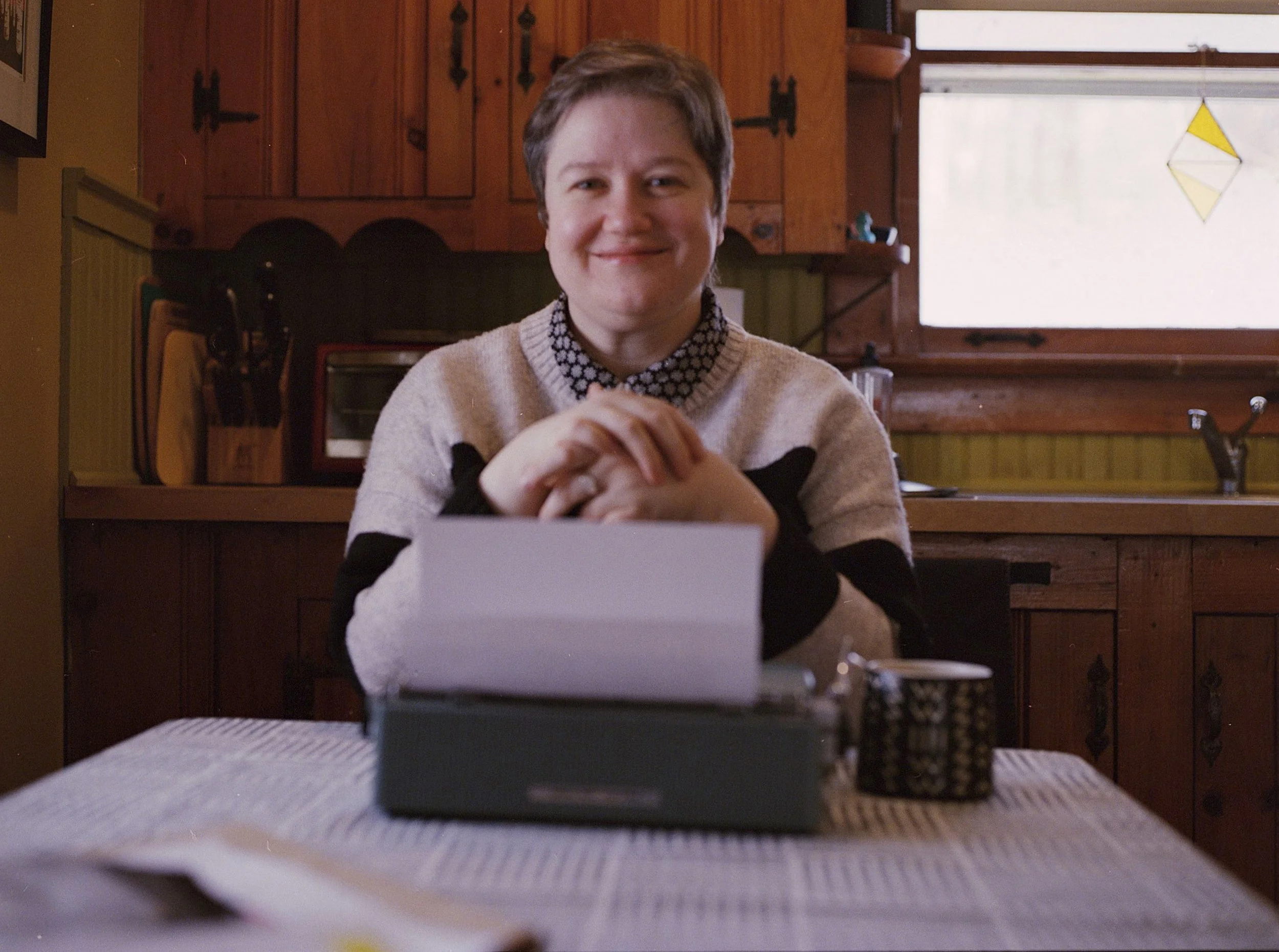 A woman sitting at a kitchen table with a smile, in front of a typewriter, wearing a beige sweater with black patterns, behind her wooden cabinets, a window with stained glass in the background.