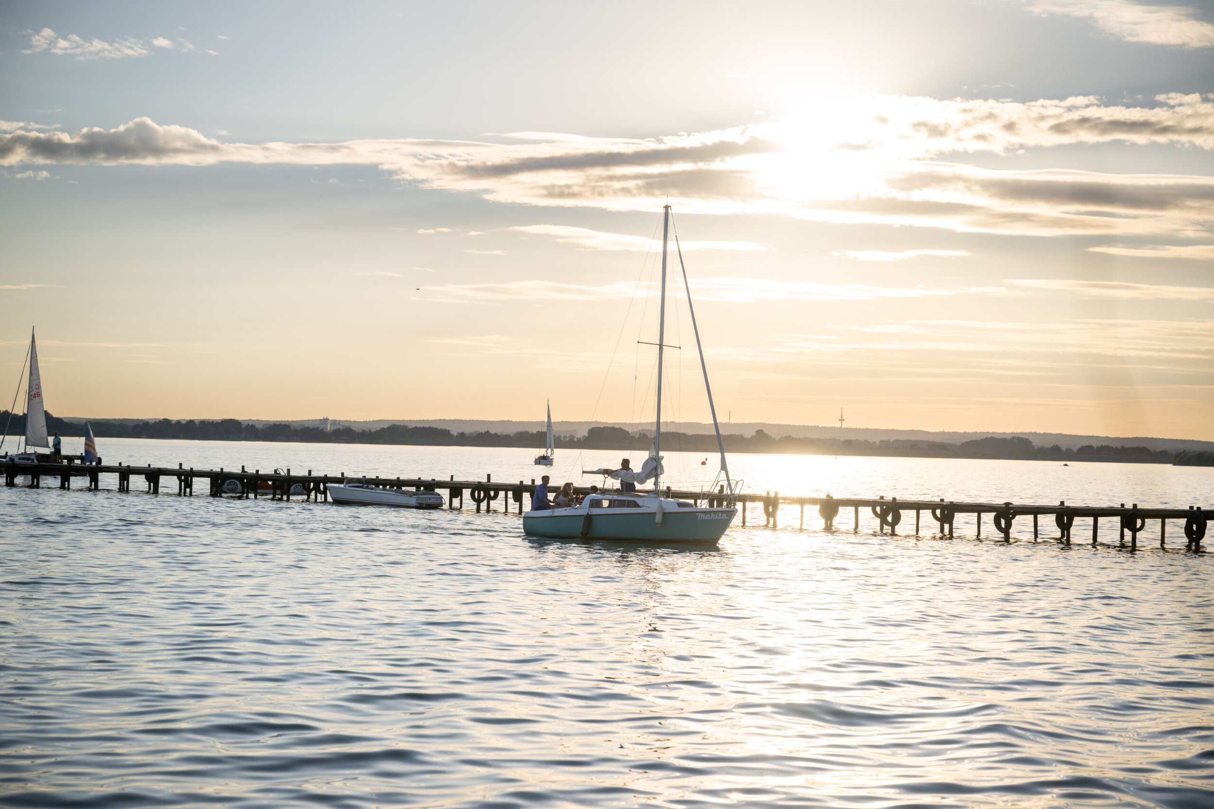 Segelboot legt am Pier bei Sonnenuntergang auf einem ruhigen See an, im Hintergrund weitere Boote und ein Himmel mit Wolken.
