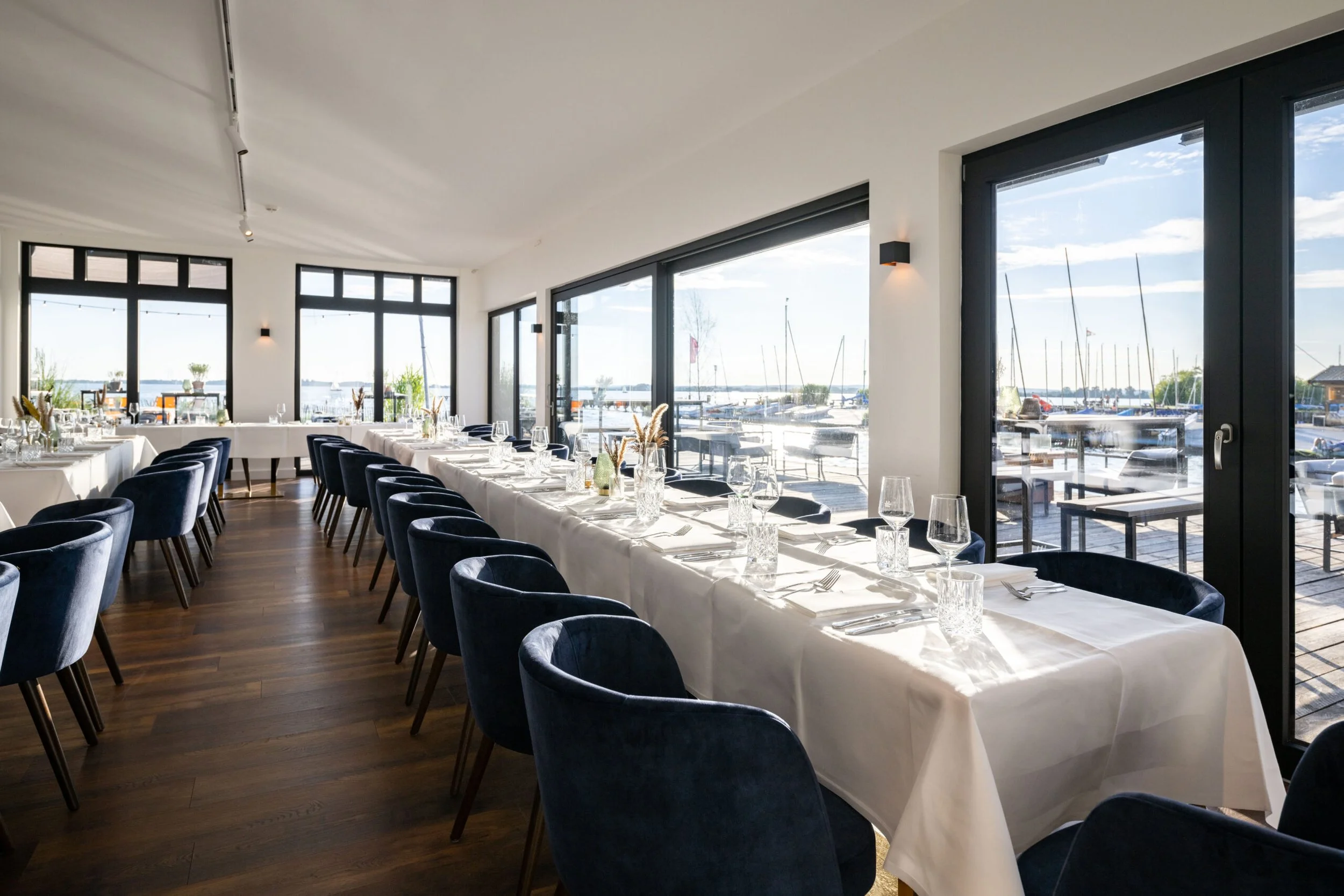 Innenraum eines Restaurants mit langem Tisch, gedeckt mit weißen Tischtüchern, Weingläsern, Wasserkrügen und Besteck, große Fenster mit Blick auf einen See mit Segelbooten.