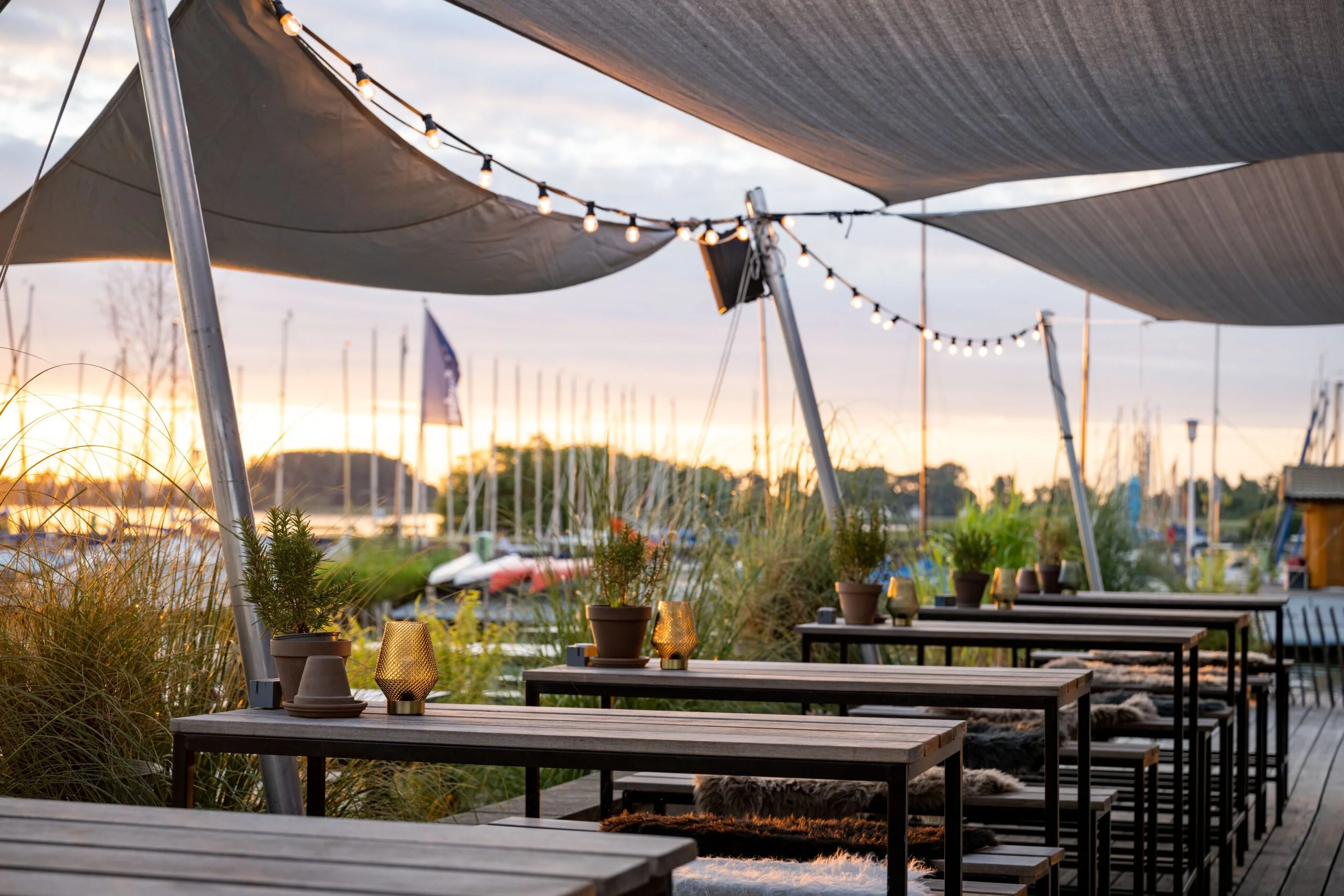 Blick auf einen Außenbereich mit Tischen und Bänken, Dekoration mit Pflanzen und Kerzen, Sonnenuntergang am Wasser mit Booten im Hintergrund, Überdachung und Lichter für eine gemütliche Atmosphäre.