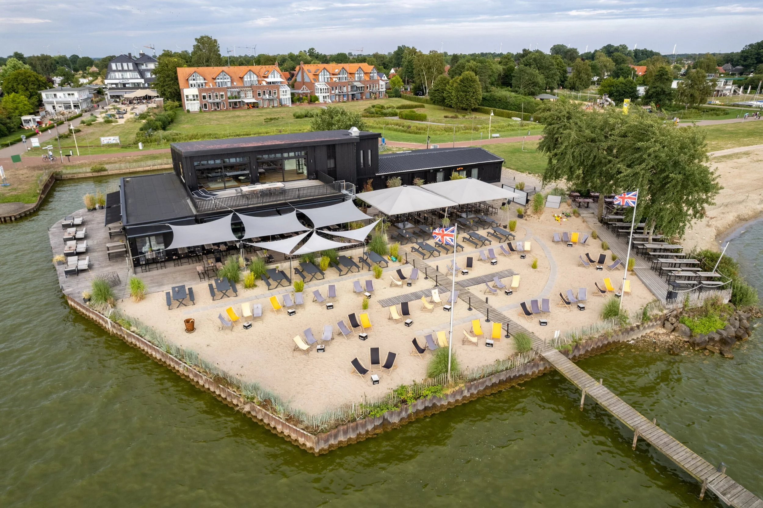 Luftaufnahme eines Strandcafés auf einer Insel im Wasser mit Liegestühlen, Sonnenschirmen und Flaggen, umgeben von Grünflächen und Wohnhäusern.