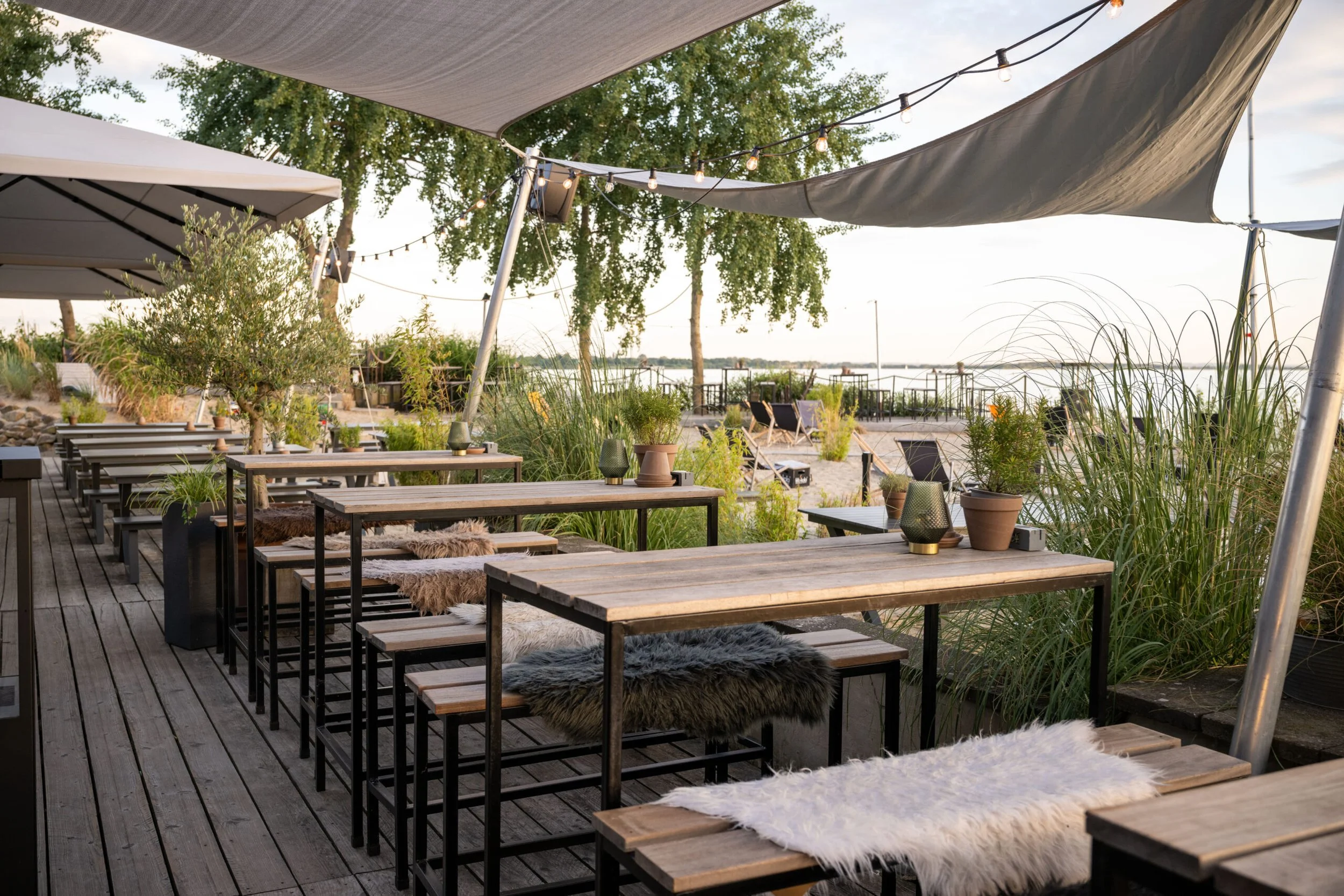 Ruhevoller Außenbereich eines Restaurants oder Cafés mit Holztischen, Stühlen und Sitzbänken, geschmückt mit Kissen aus Fell und Pflanzen, unter einem großen Sonnensegel und Lichterketten, mit Blick auf einen Wasserarm mit Ufer und Möwen im Hintergru