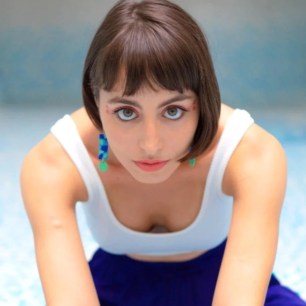 Jeune femme avec des cheveux bruns courts, portant un débardeur blanc et de grandes boucles d'oreilles bleues et vertes, regardant directement l'objectif.