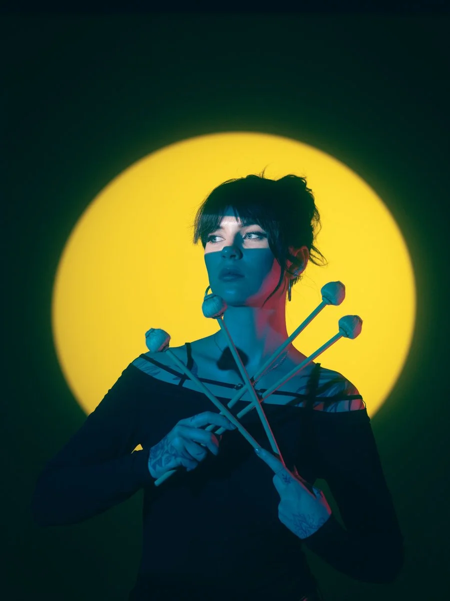 Une femme aux cheveux courts, portant un vêtement noir à rayures, se tient devant un fond jaune lumineux, tenant quatre maillons de métal avec des bolas à chaque extrémité, et regarde à gauche.