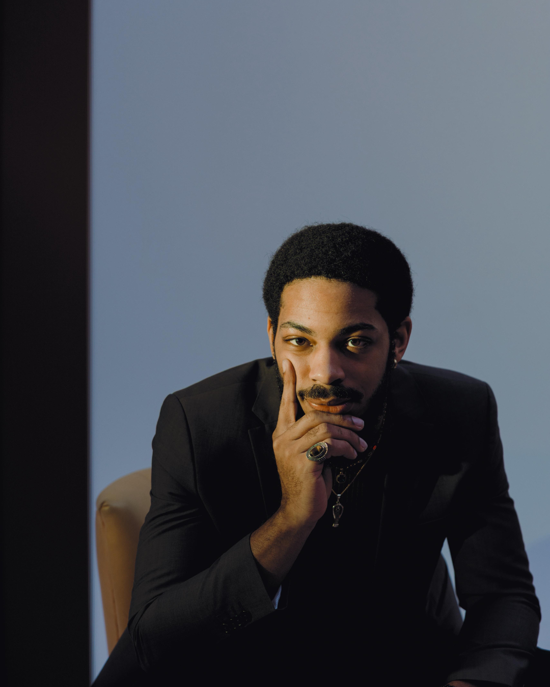 Un homme afro-américain en costume noir, assis à une table, regardant pensivement vers l'appareil photo avec la main sur son visage.