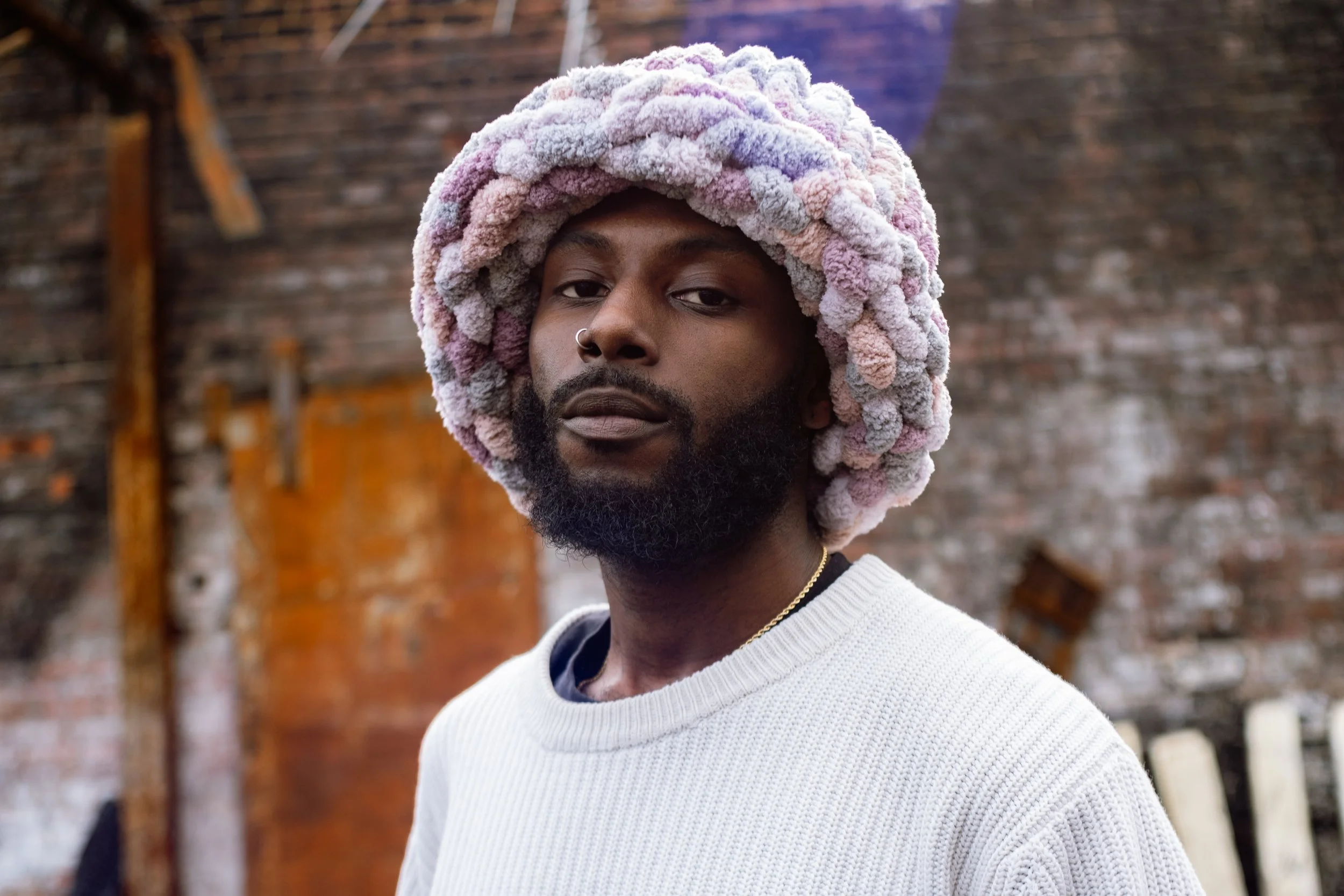 Un homme portant un bonnet volumineux en peluches de couleurs pastel, portant un pull blanc, dans un environnement urbain avec un mur en briques en arrière-plan.