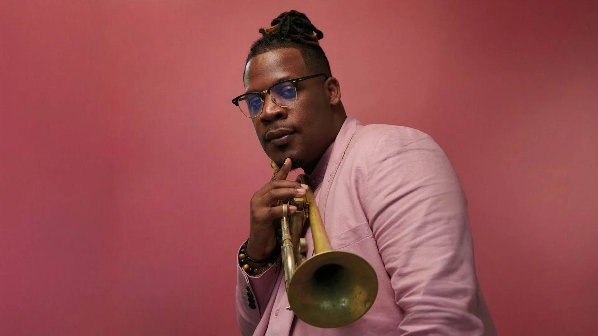 Un homme portant des lunettes, une veste rose et des bracelets tient une trompette, posé contre un fond rose.