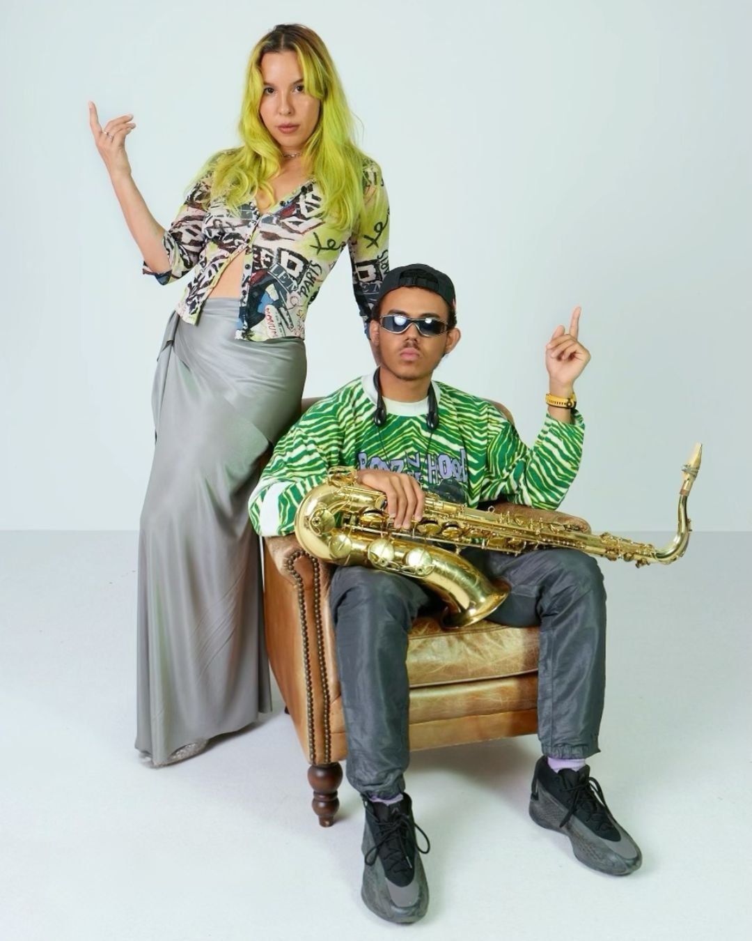 Deux jeunes, un homme assis avec un saxophone et une femme debout à côté, posant dans un studio avec un fond blanc.
