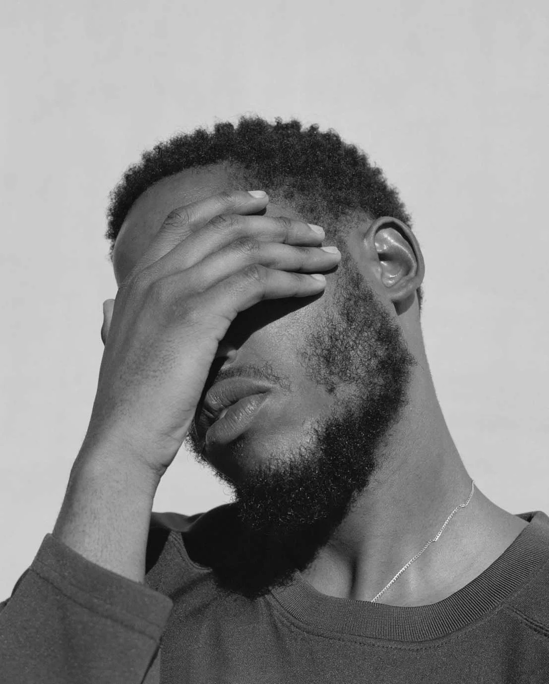 Un homme noir porte sa main sur son visage, couvrant ses yeux, avec une expression de réflexion ou de gêne, en noir et blanc.