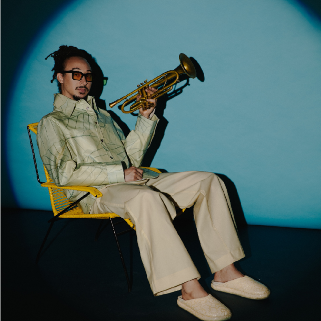 Un homme assis sur une chaise jaune, tenant un trompette, dans un espace sombre avec un fond bleu clair
