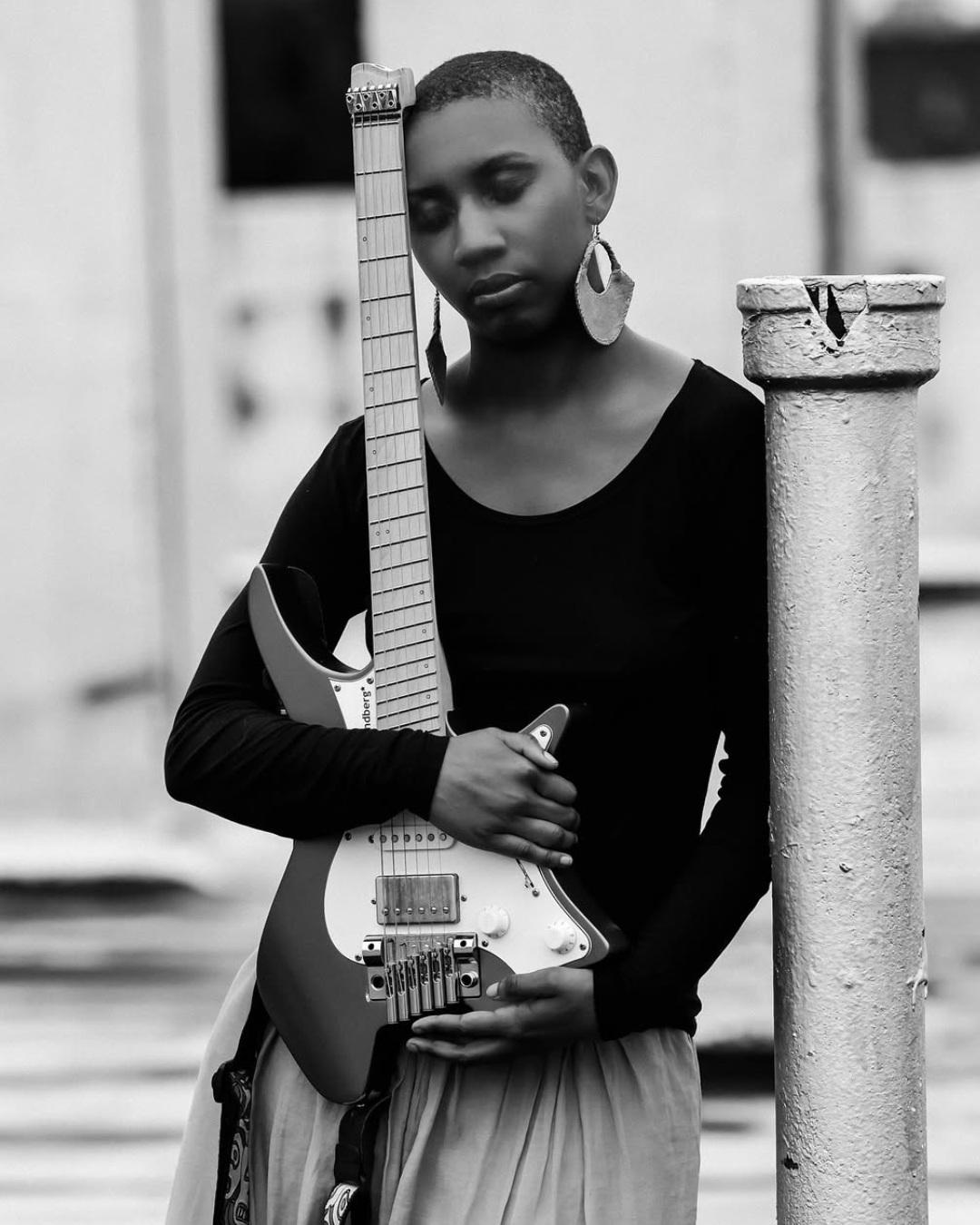 Une jeune femme avec une coupe de cheveux courte, portant de grandes boucles d'oreilles, tenant une guitare électrique dans une ambiance urbaine en noir et blanc.