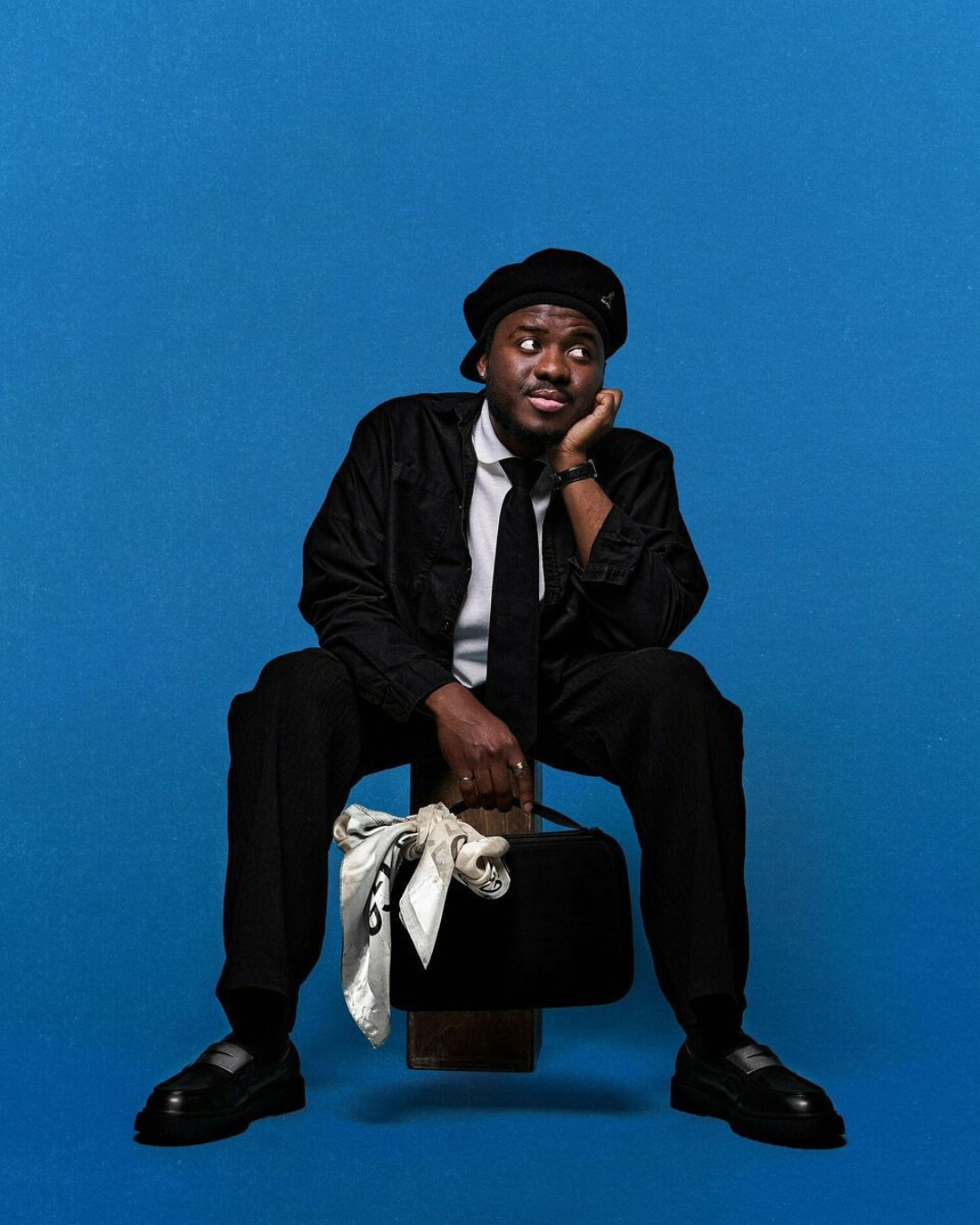 Jeune homme noir assis sur un tabouret, portant des vêtements noirs, une chemise blanche, une cravate noire, un béret noir, avec un sac noir et un foulard blanc à la main, contre un fond bleu.