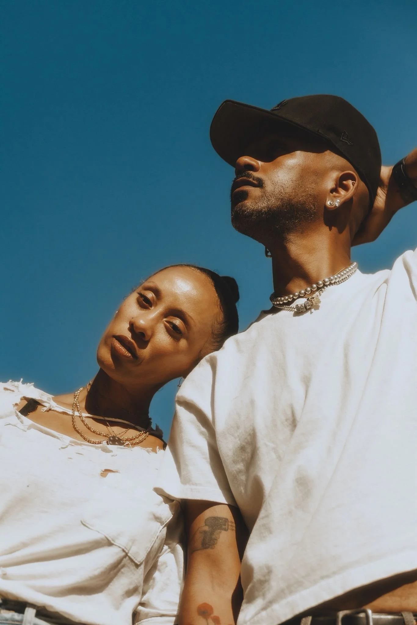 Deux personnes posant sous un ciel clair, portant des vêtements décontractés, avec des bijoux, l'une portant une casquette et l'autre affichant une expression confiante.