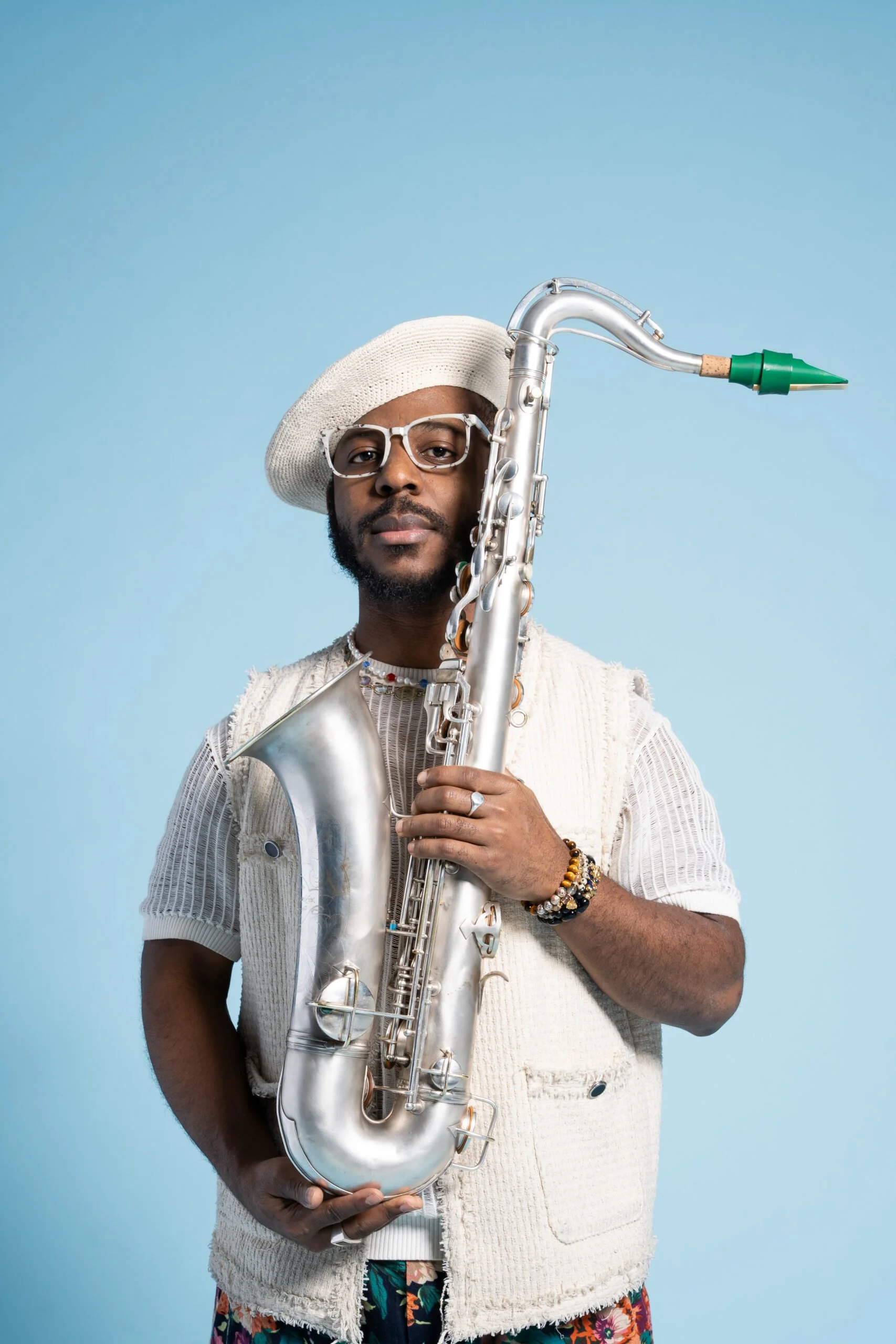 Un homme portant des lunettes et un chapeau blanc, debout contre un fond bleu clair, tenant un saxophone d'argent.