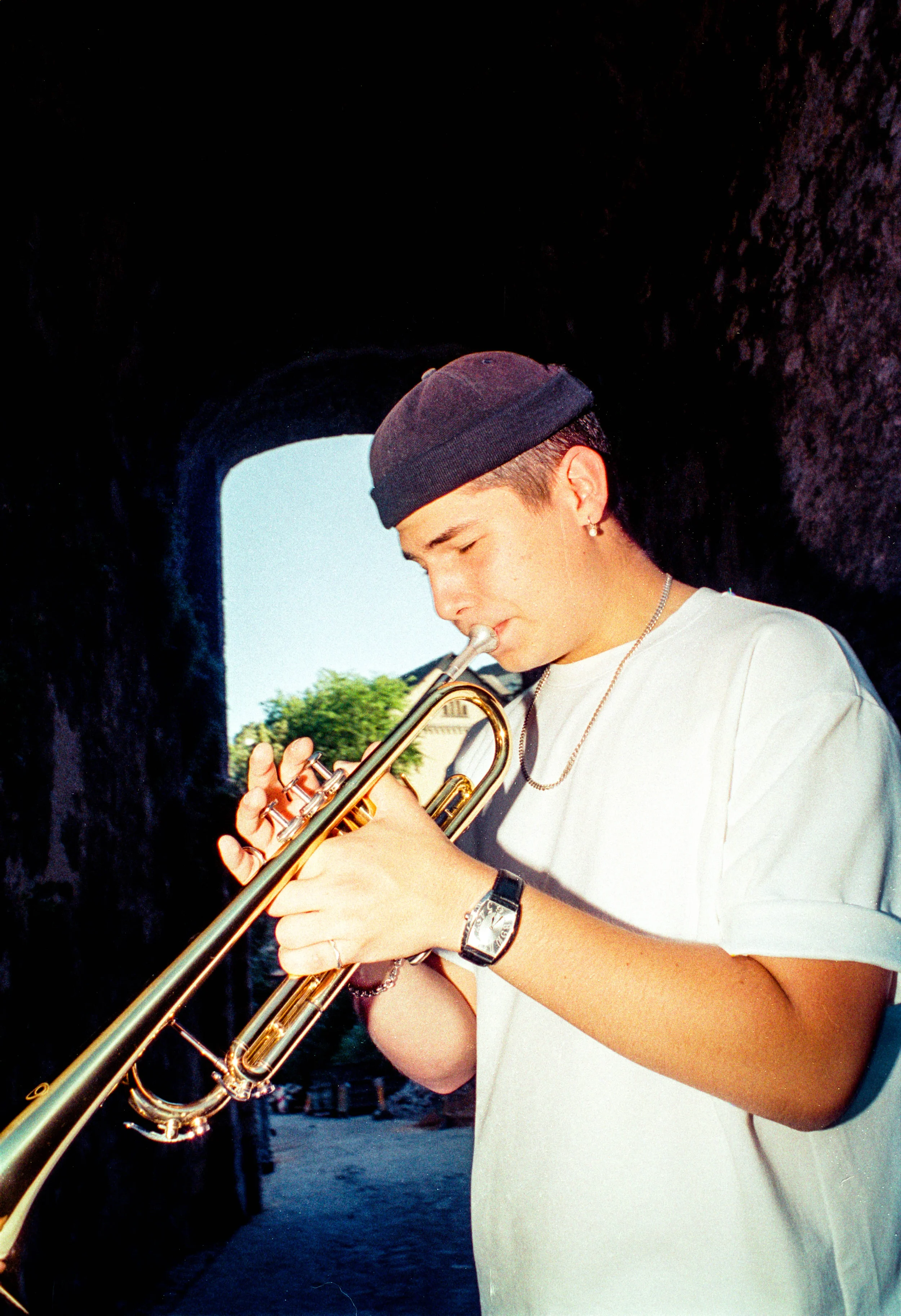Jeune homme jouant de la trompette, portant un bonnet noir, une chemise blanche, dans un tunnel avec un ciel clair et des arbres en arrière-plan.