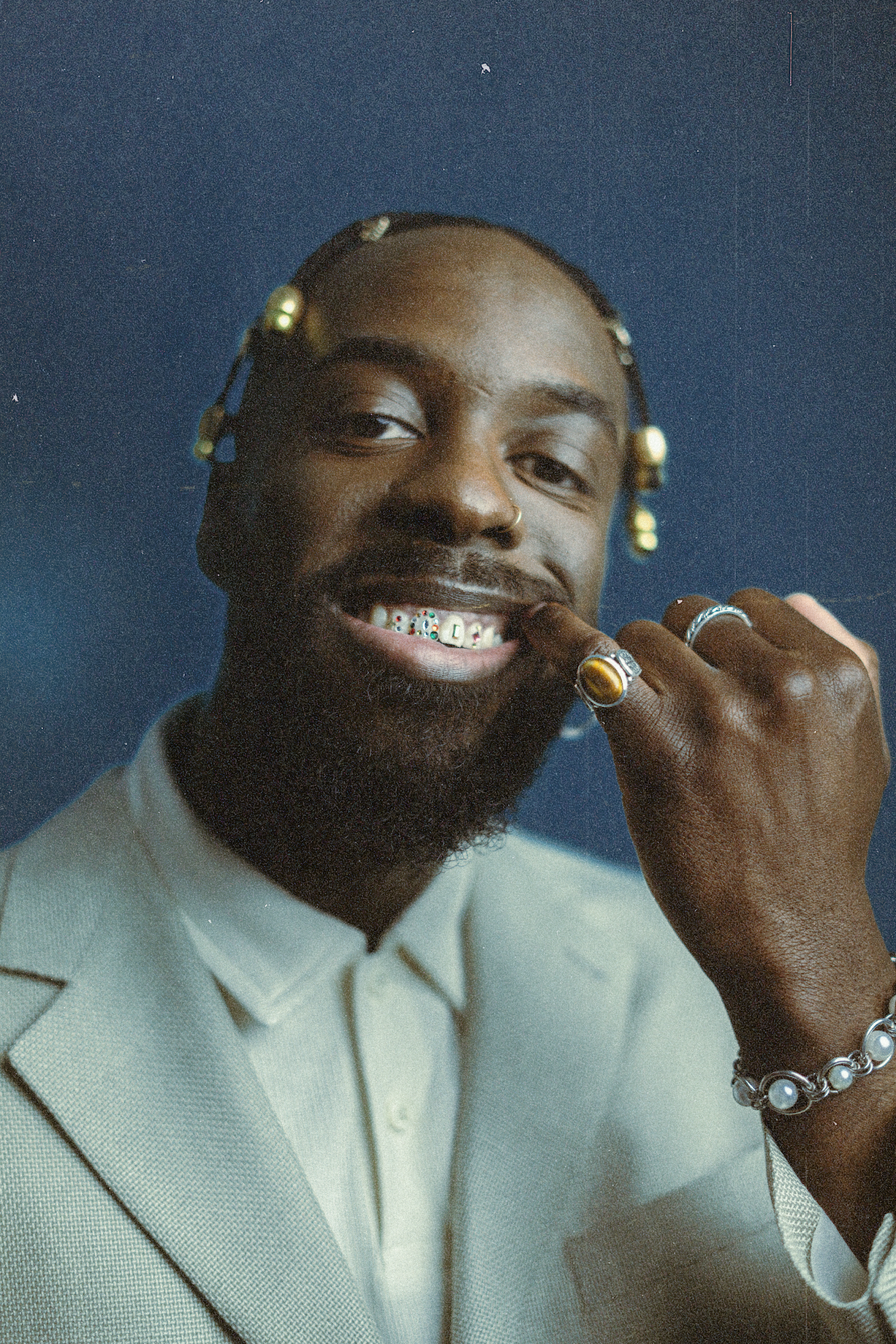 Portrait d'un homme noir souriant avec des dents de dents colorées, portant des bijoux et un costume clair, contre un fond bleu.