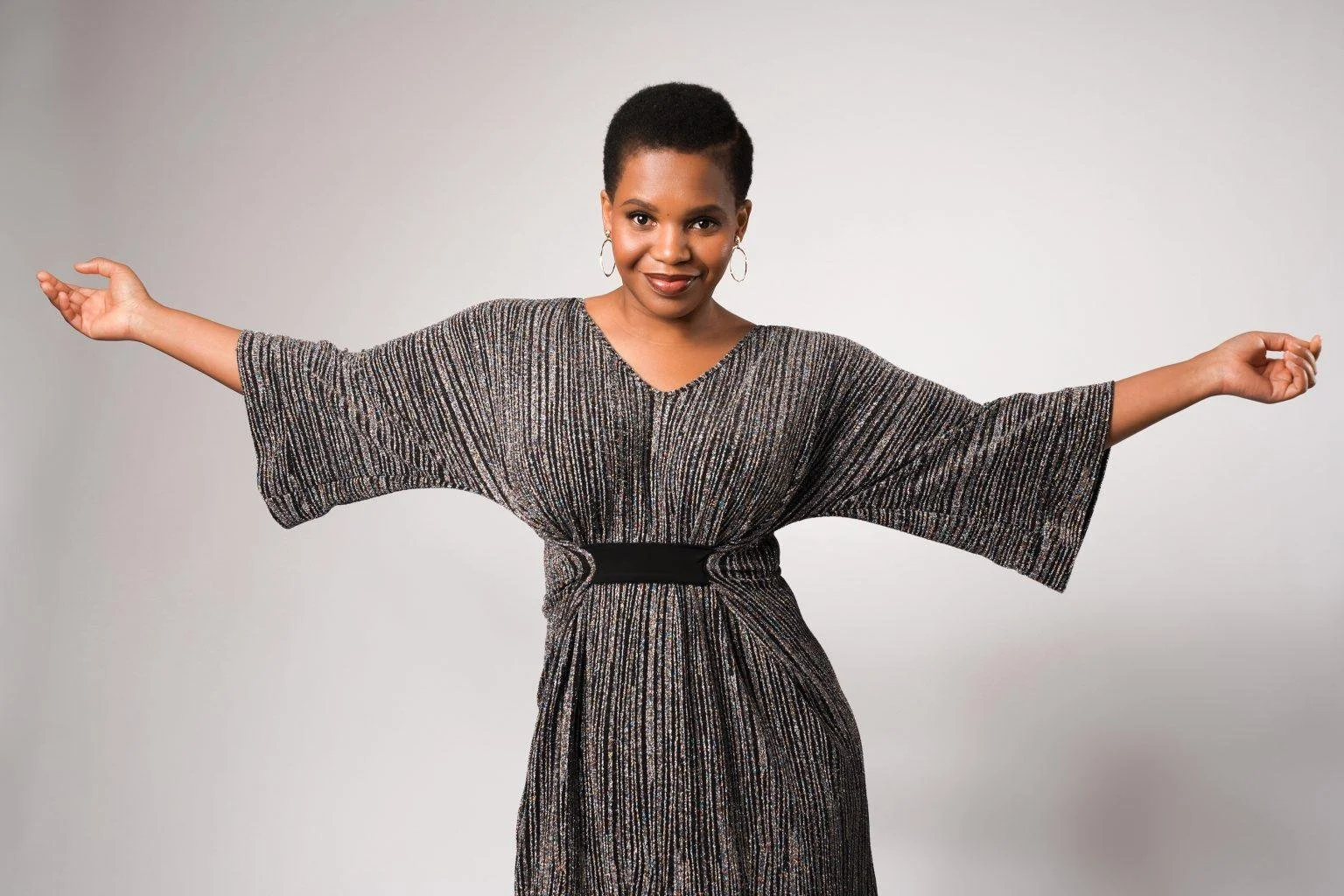 Une femme souriante avec des bras ouverts, portant une robe à manches larges en tissu brillant, fond gris clair.
