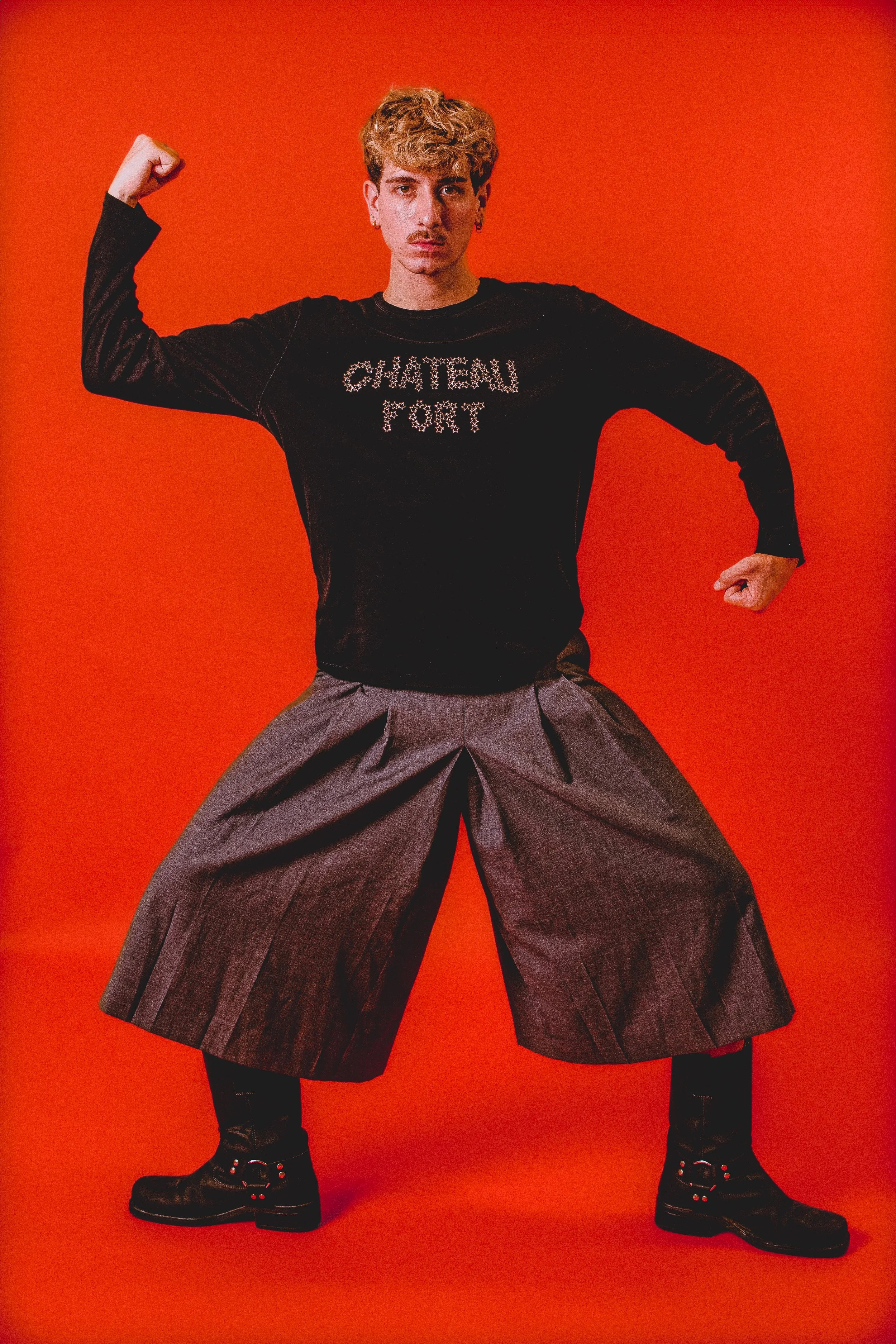 Jeune homme posant en flexant ses bras, portant un pull noir avec 'Chateau Fort' écrit dessus, des pantalons larges gris et des bottes noires, sur un fond orange.