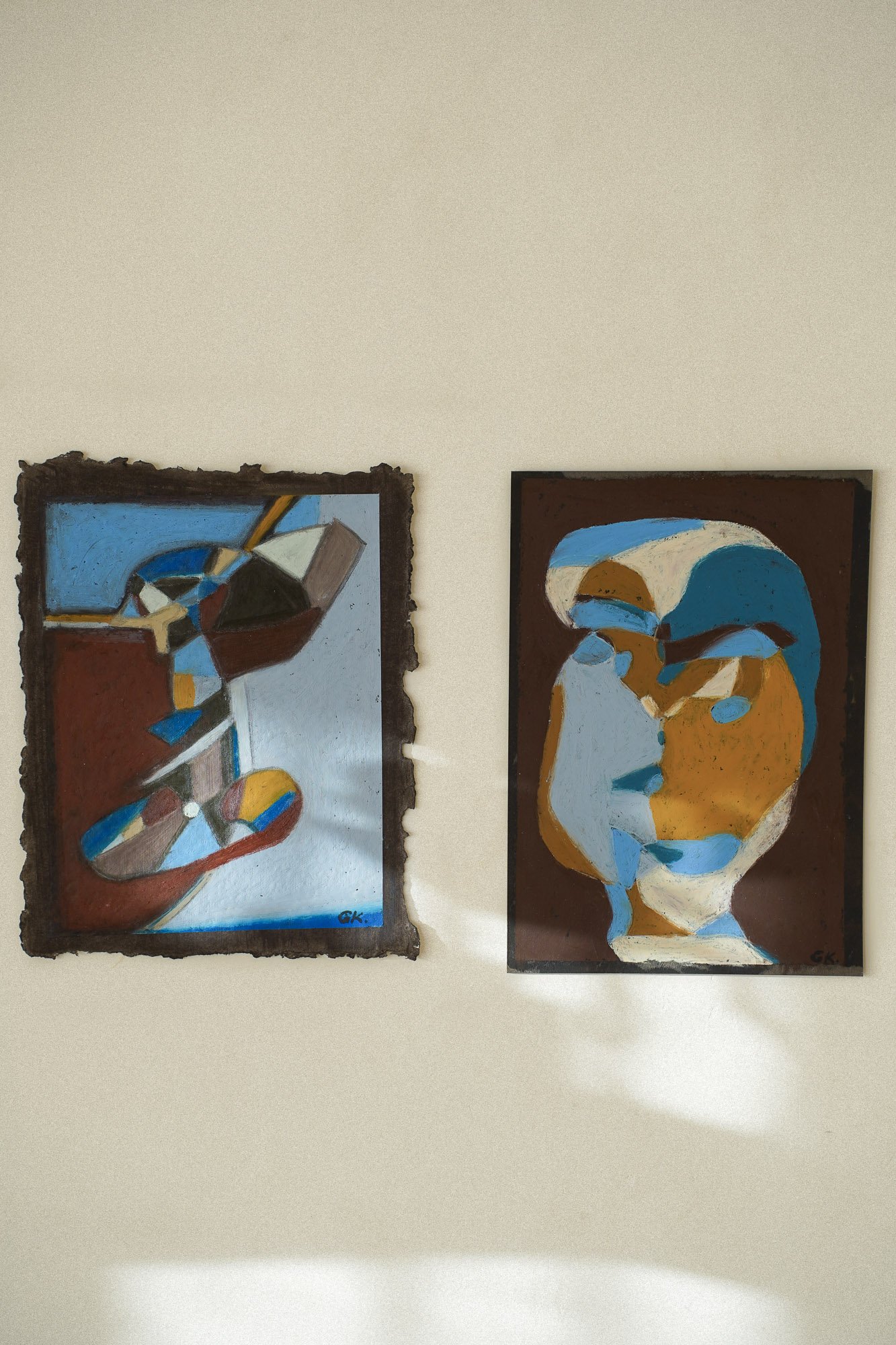 Two figurative abstract paintings hung on a white wall, in an art studio in Oslo. The left painting features geometric shapes and muted colors, while the right painting depicts a cubist-inspired human face with bold, contrasting colors.