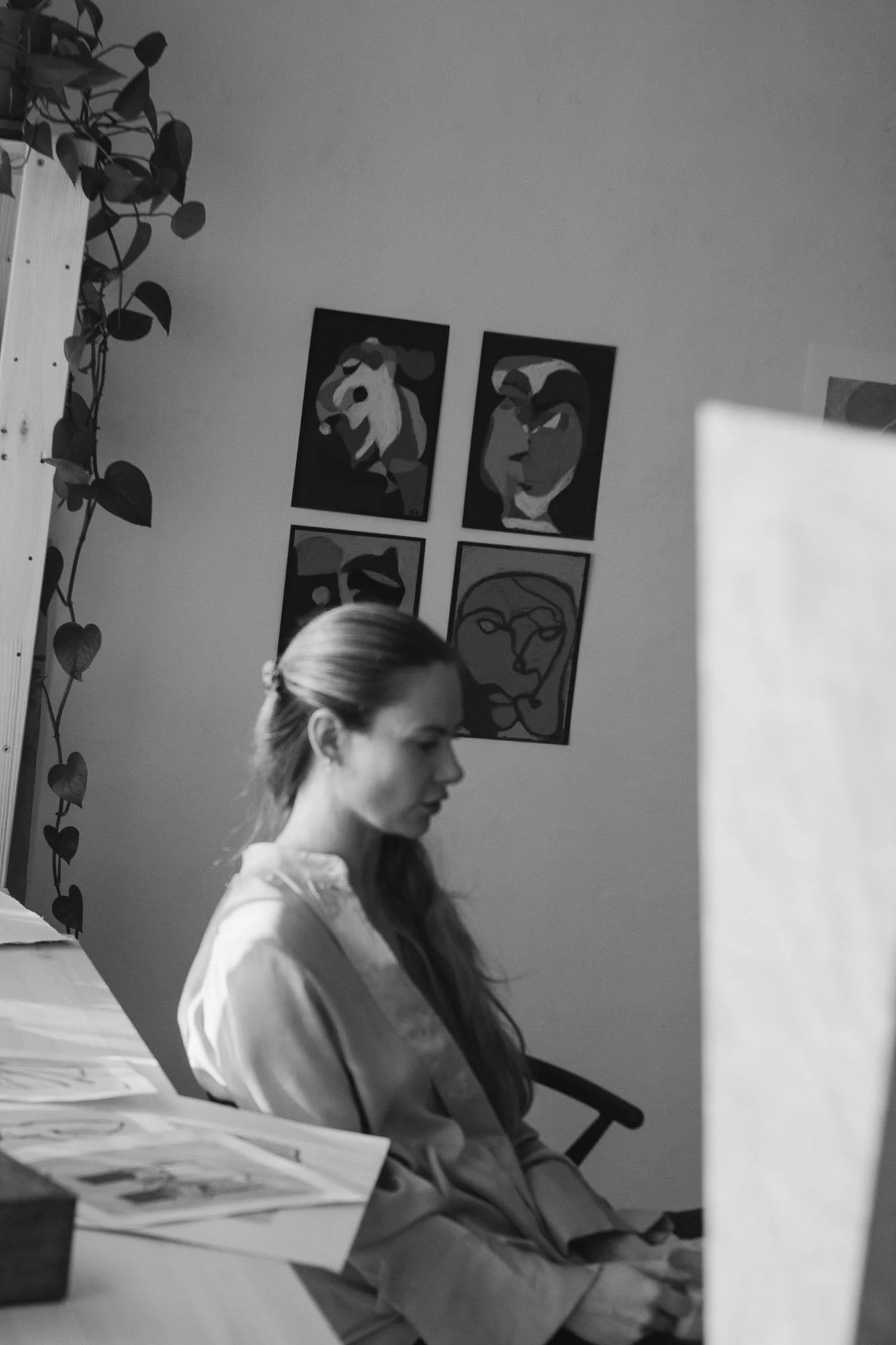 A woman, Norwegian artist, sitting on a chair with her eyes closed, in a room with four abstract, figurative portrait paintings on the wall behind her, a table with papers, unfinished artwork and a plant on the left side.