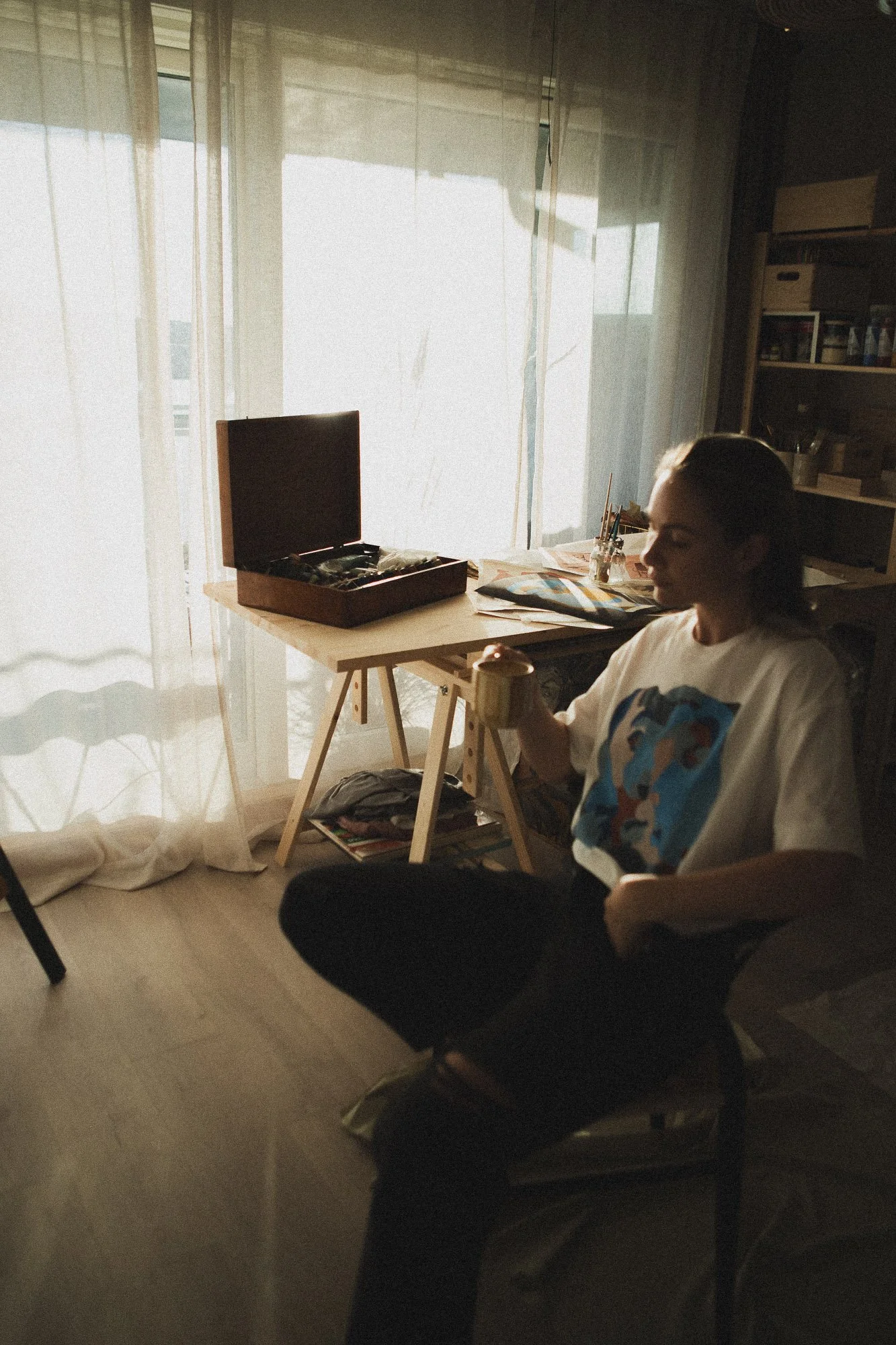 A woman, Norwegian artist is sitting in a chair in a dimly lit room in her art studio in Oslo. With sunlight coming through curtains, holding a mug of coffee, and looking down.