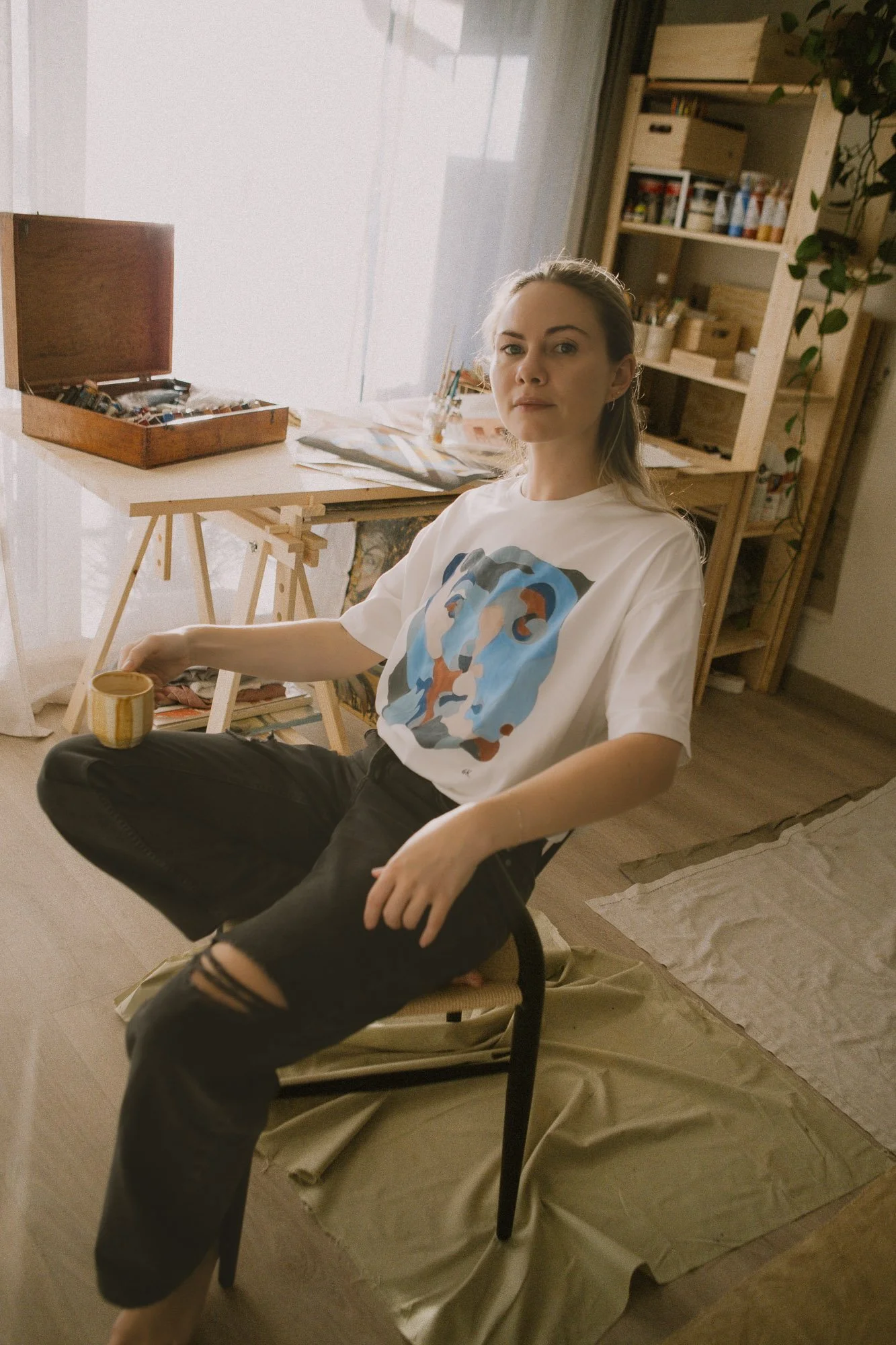 A young woman, Norwegian artist, wearing a white t-shirt with a colorful abstract face print and black ripped jeans, is sitting on a chair in an art studio, holding a cup, with shelves, art supplies, and a work table in the background.