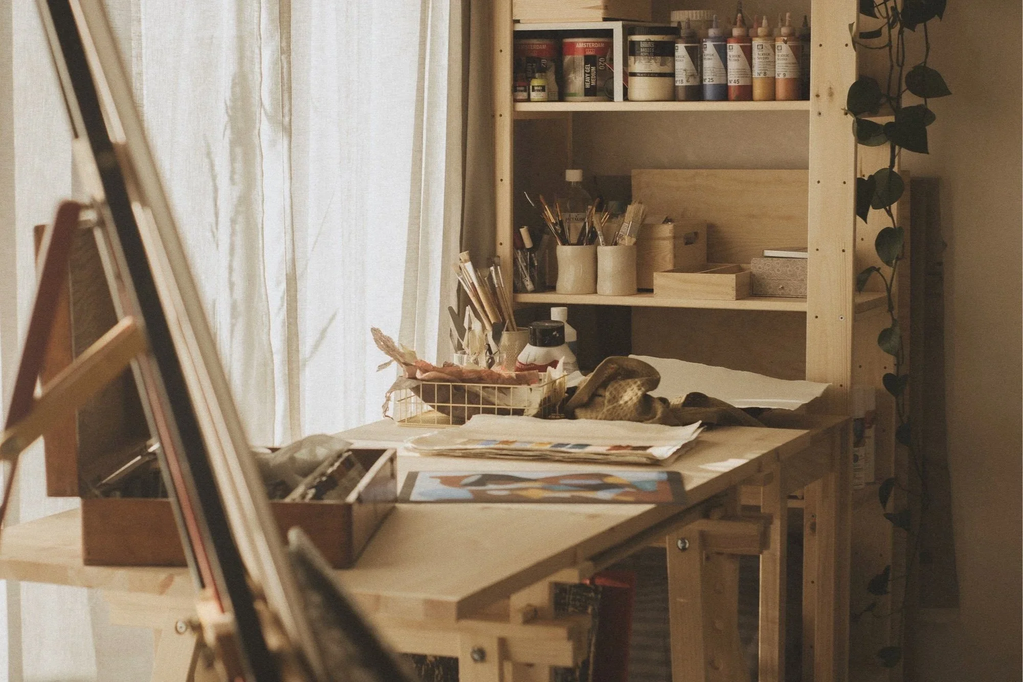 An art studio in Norway. A wooden art and craft workspace with shelves holding paints, brushes, and boxes, featuring a table with art supplies, papers, and a small wire basket containing various items.