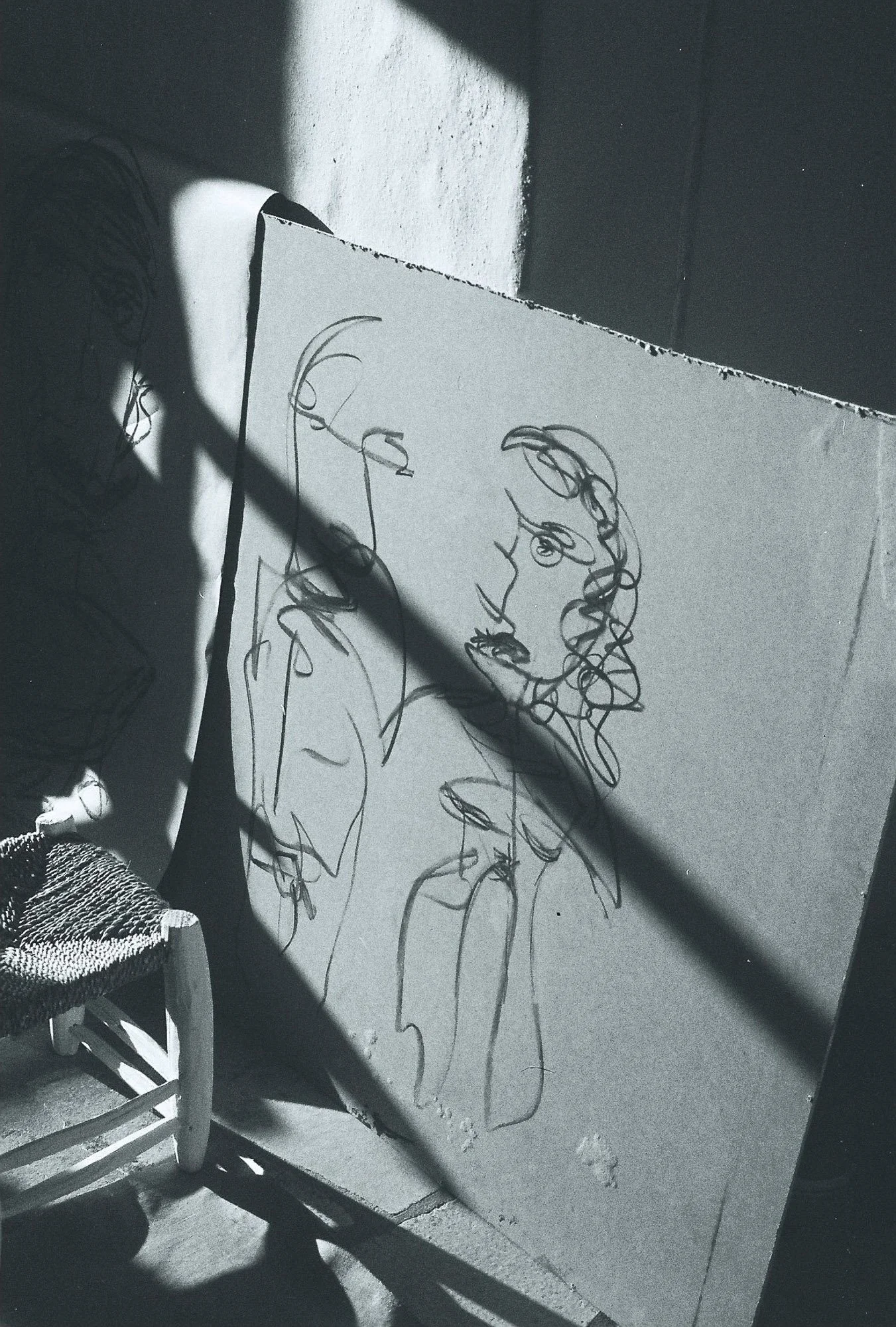 Black and white photo of an abstract line art portrait of a woman on a large canvas, with a shadow crossing the face. A woven bag is visible in the lower left corner.