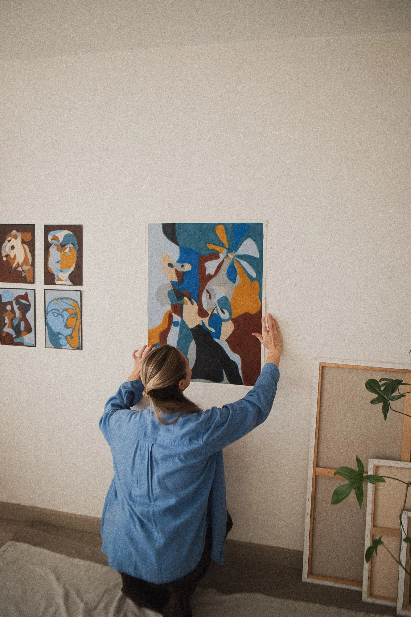 An Norwegian artist with blonde hair in a ponytail wearing a blue shirt is hanging an expressive abstract, figurative painting on a white wall in an art studio in Oslo, with several other paintings and artistic materials visible in the background.