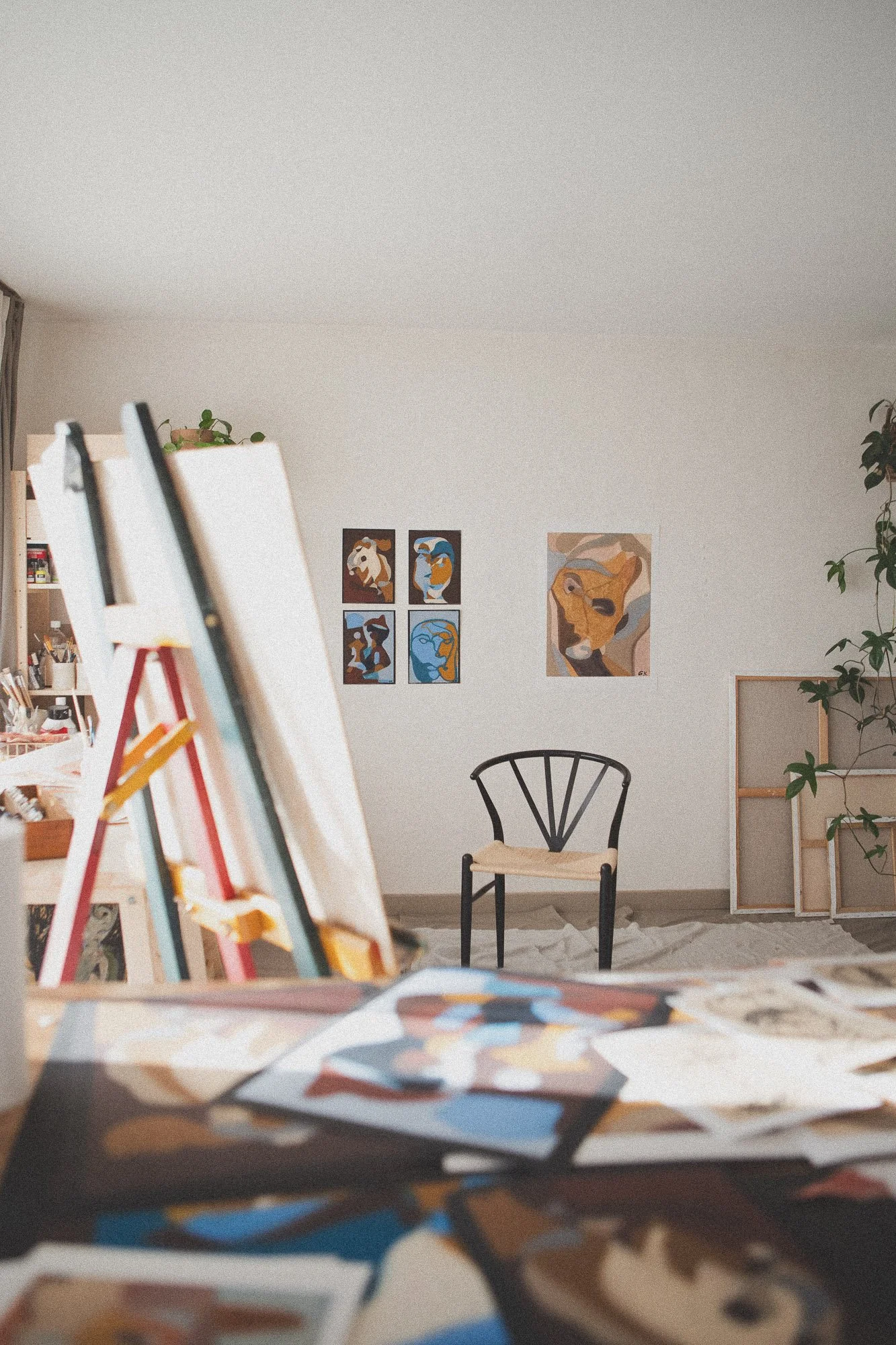 An art studio in Oslo with expressive figurative abstract paintings on easels and unfinished artwork, a black chair against a white wall decorated with abstract portraits, and a tall green plant in the corner.