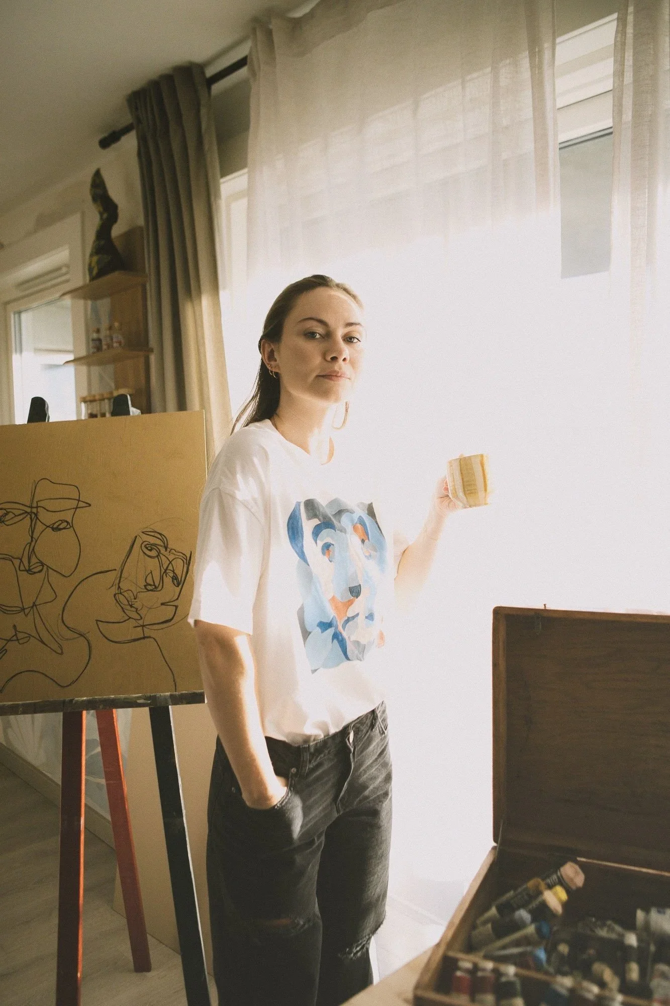 A woman standing in an art studio, holding a sponge, in front of a window with sheer curtains. She wears a white t-shirt with a dog illustration, with her left hand in her pocket. There is an easel with a line drawing of faces and an open wooden box with art supplies on a table.