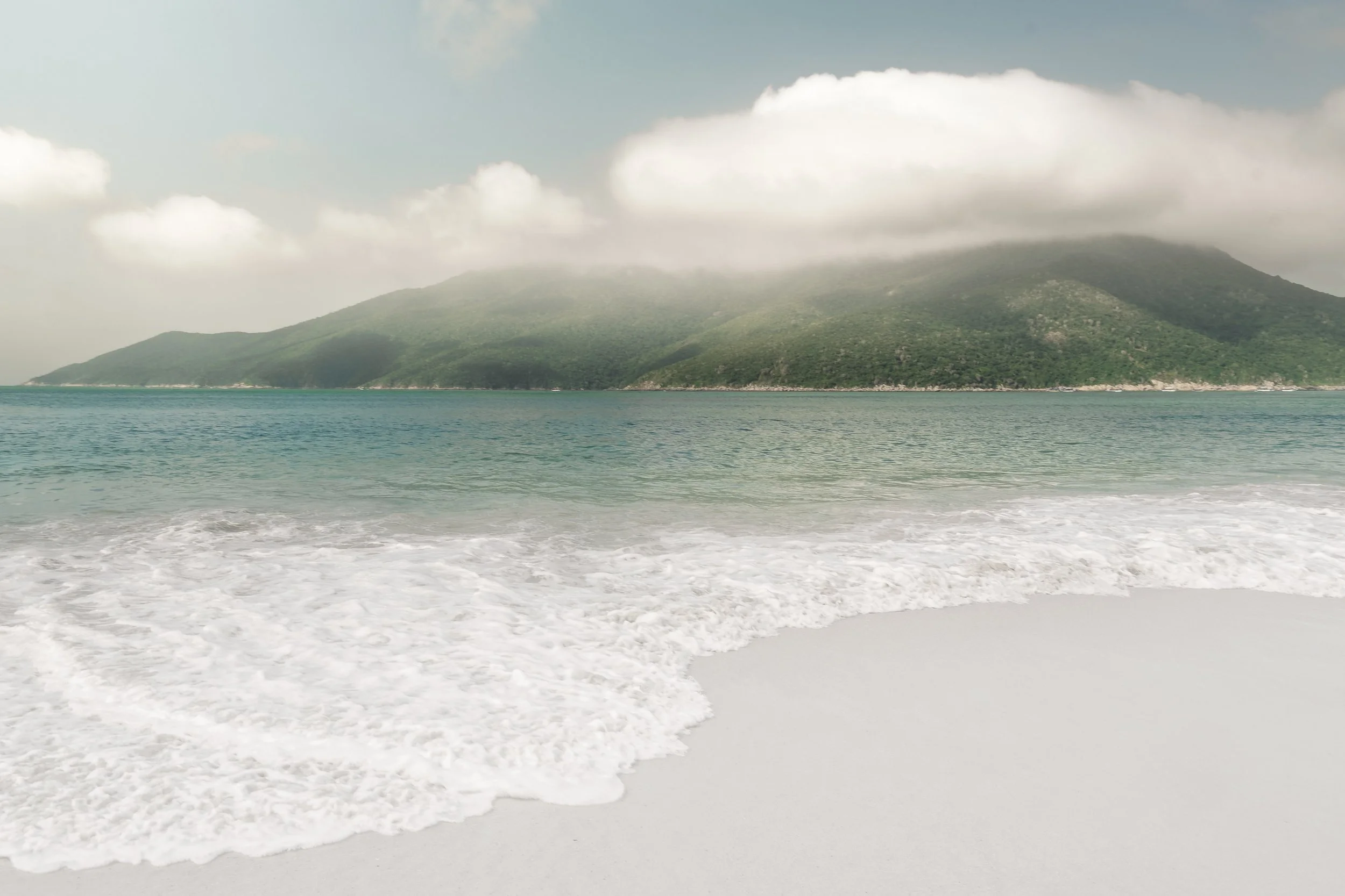 Onda branca chegando na areia com serra verde ao fundo, focada em decoração residencial e corporativa que usa natureza como ponto visual principal.
