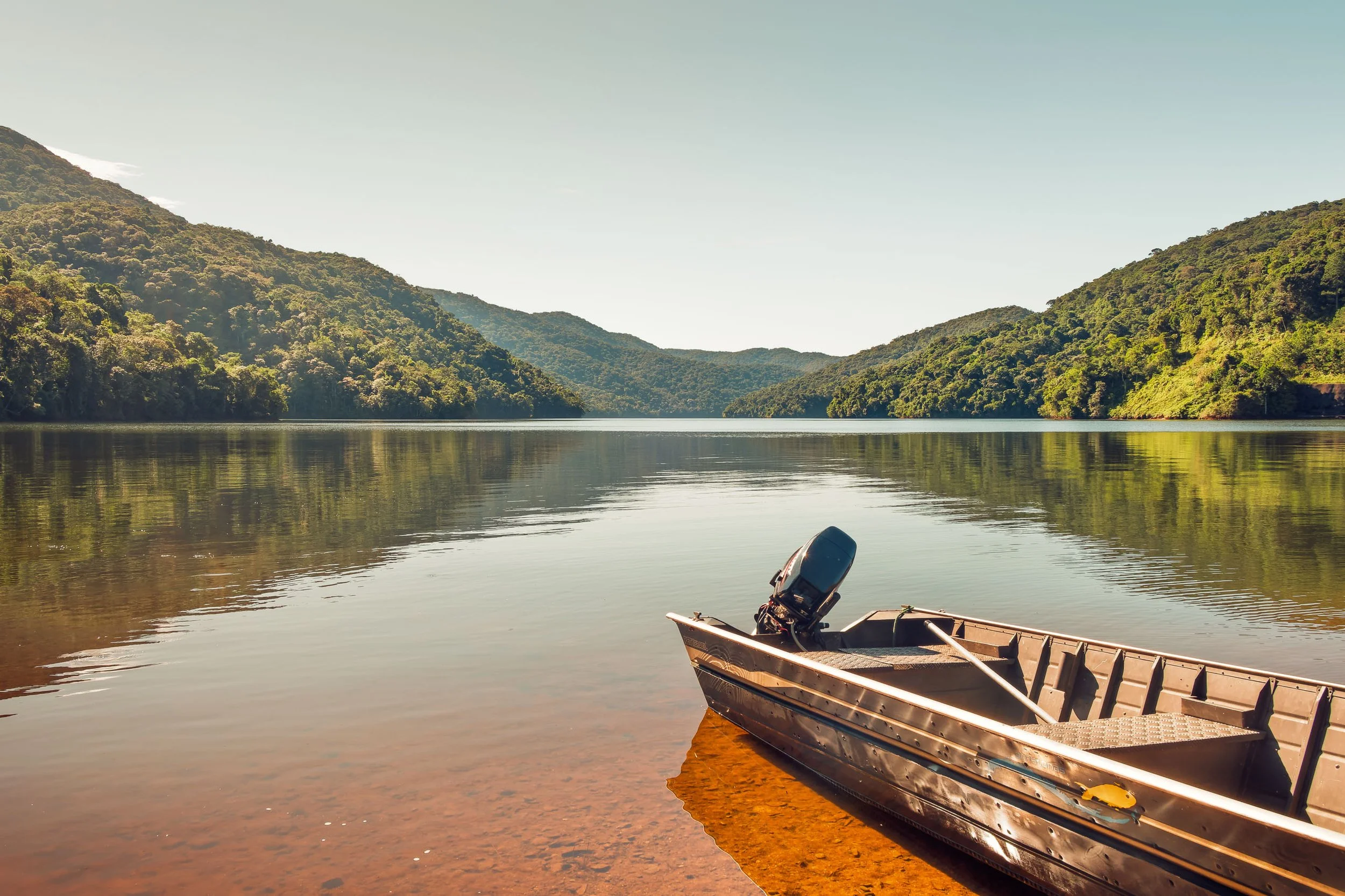 Barco metálico parado na margem da represa do Legado das Águas com montanhas verdes refletindo na água calma. Céu limpo e paisagem silenciosa. Foto indicada para decoração com foco em natureza brasileira e sensação de tranquilidade.
