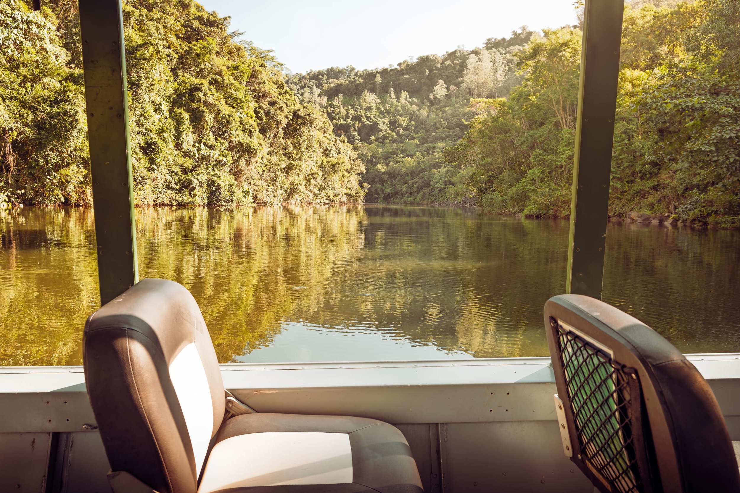 Vista da represa do Legado das Águas através da janela de um barco com assentos vazios. Água calma refletindo a mata densa. Fotografia sem pessoas, indicada para interiores que buscam sensação de descanso e paisagem brasileira preservada.
