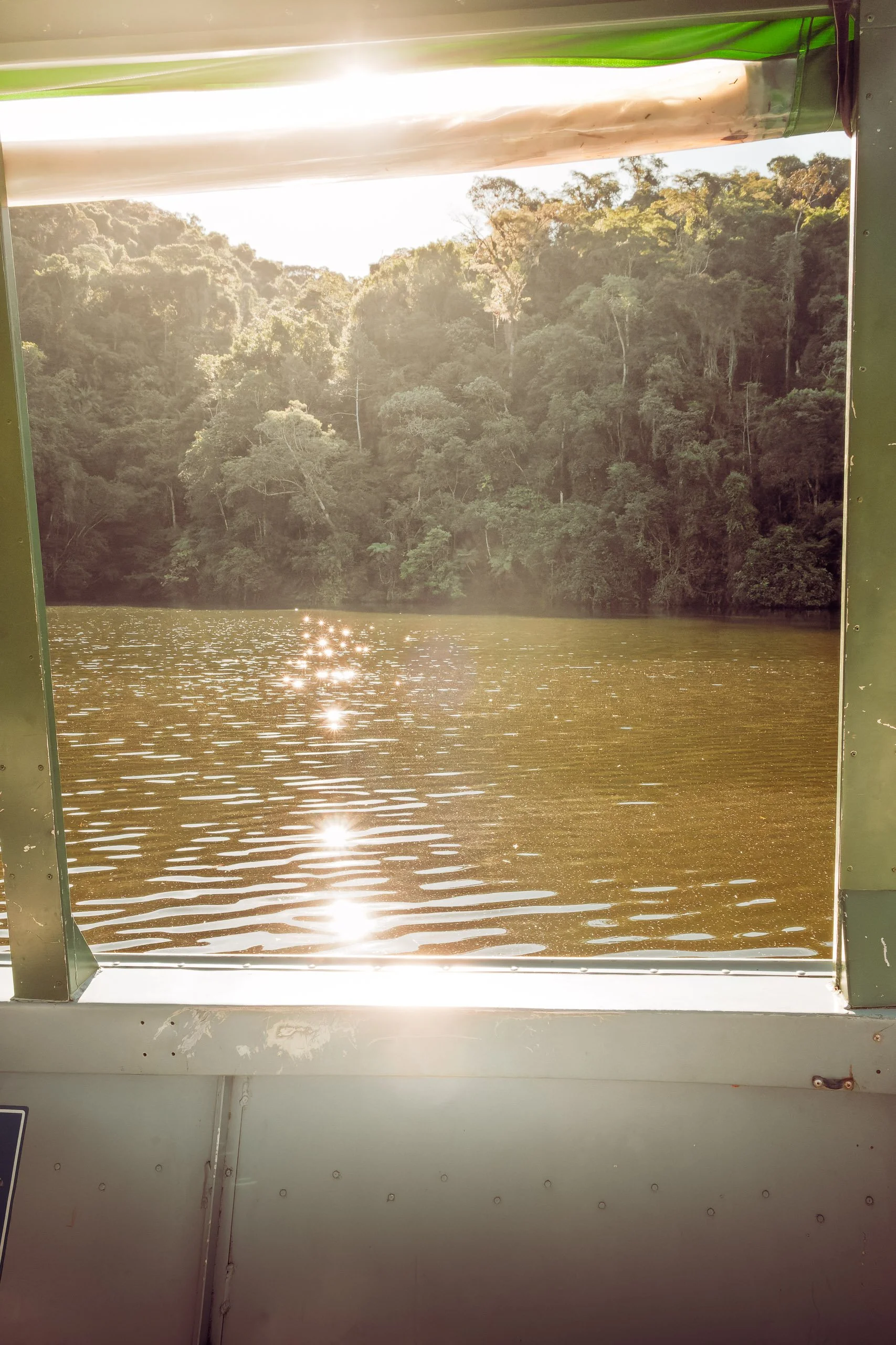 Reflexos de sol sobre a superfície da represa do Legado das Águas vistos de dentro de um barco. Mata densa ao fundo e brilho quente no horizonte. Fotografia pensada para ambientes que pedem sensação de luz e serenidade.
